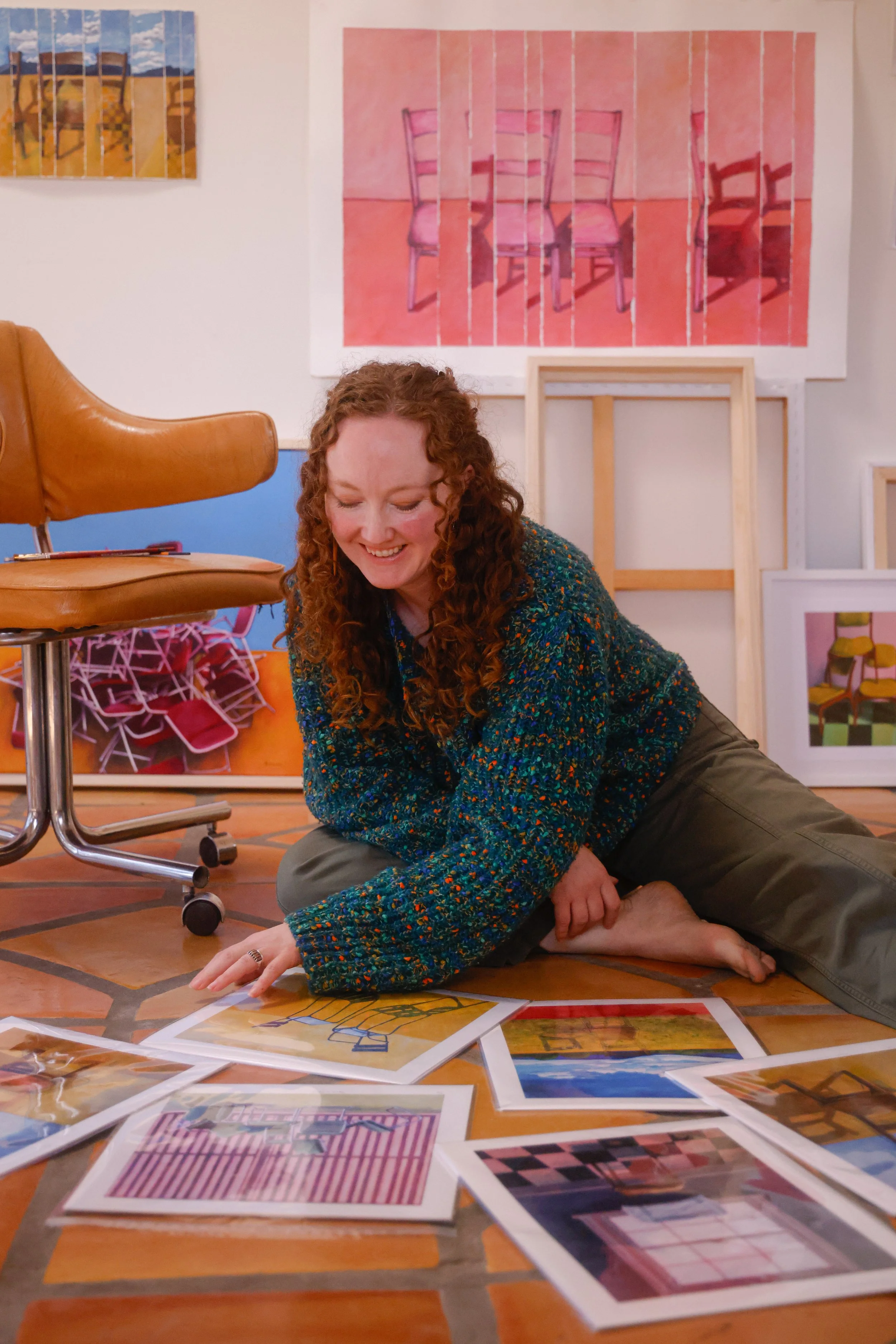 Mujer con cabello rizado y suelto, vestida con un suéter de colores y pantalón caqui, sonriendo y buscando en varias fotografías de arte que están dispersas en el suelo de un estudio de arte con cuadros y lienzos en el fondo.