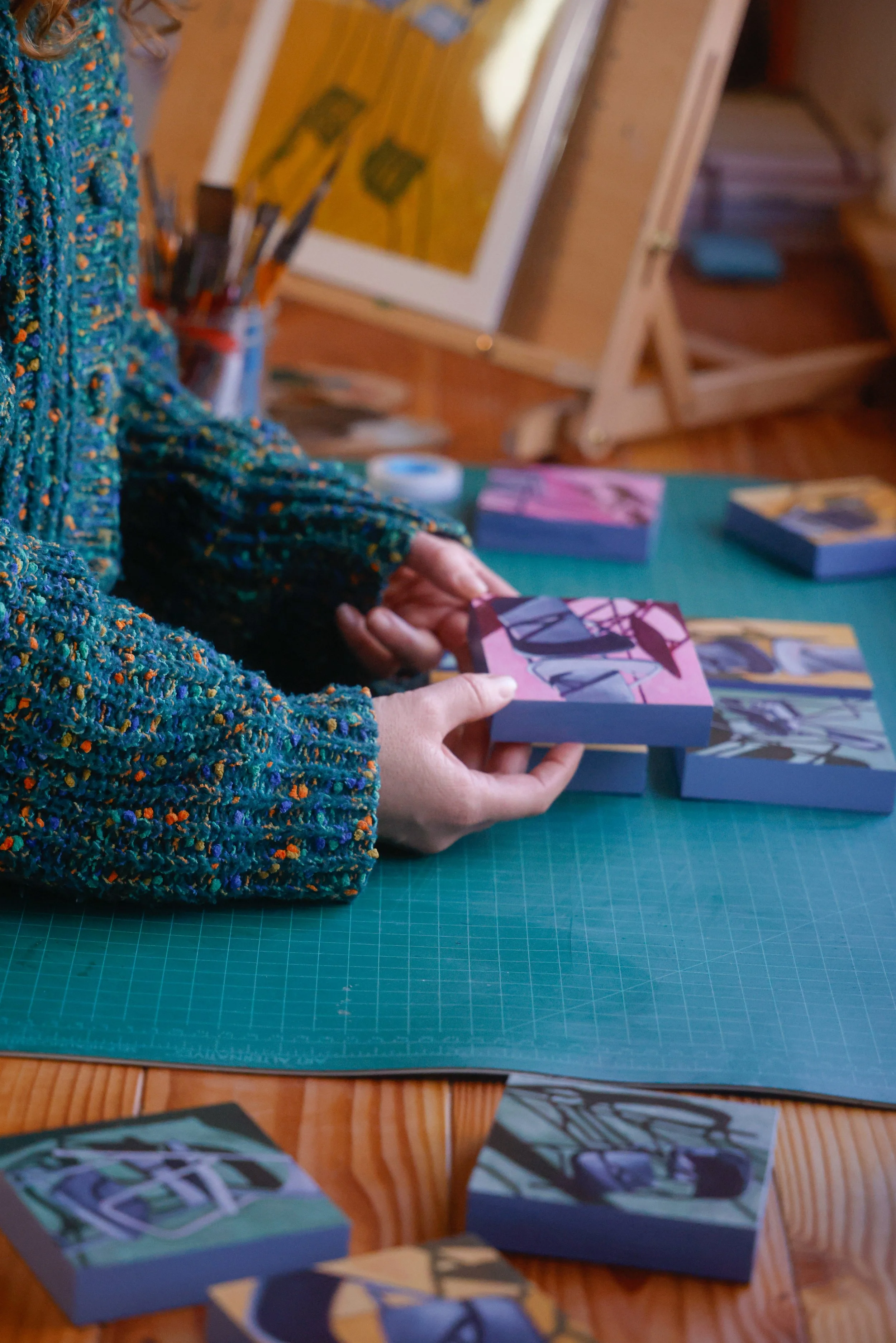 Una persona sosteniendo un pequeño cuadro con arte abstracto en una mesa verde, rodeada de otros cuadros en miniatura y materiales de arte, en un taller o estudio.