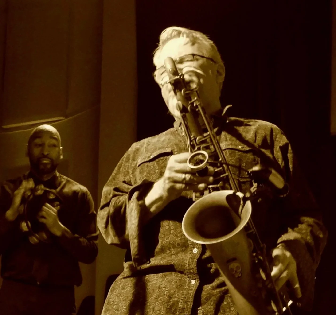Two musicians, one playing a saxophone and the other holding a percussion instrument, performing on stage in a dimly lit setting.