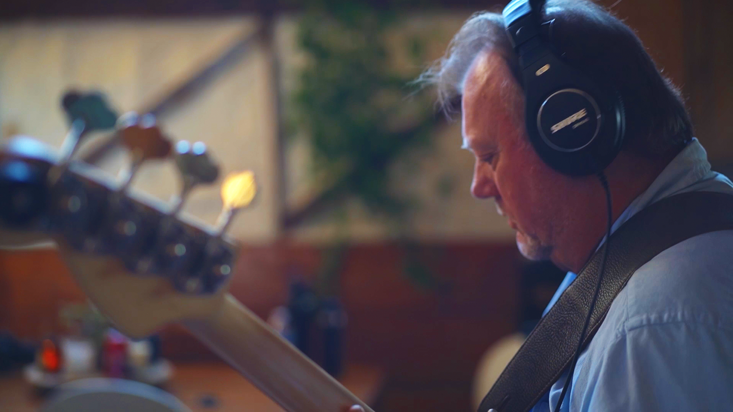 A man with gray hair and a beard wearing headphones and holding a guitar, focusing on the guitar neck in a warm, softly-lit room.