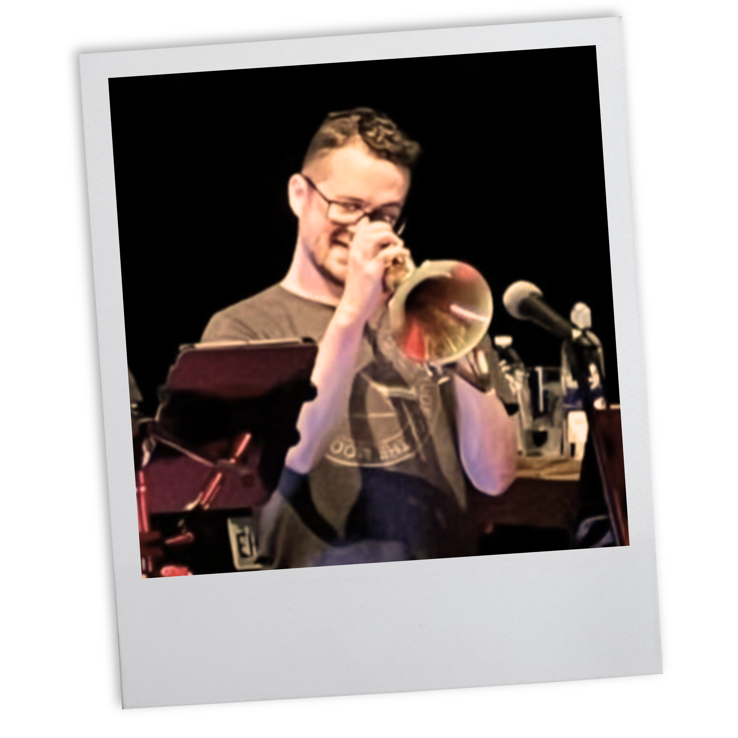 A man wearing glasses and a dark t-shirt plays a trumpet on stage, with a microphone and various drinks in the background, appearing to be performing or practicing.