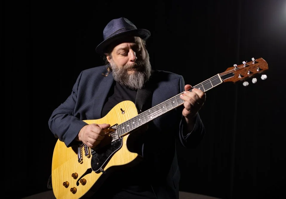 A man with a beard, wearing a fedora hat and a dark jacket, playing an electric guitar on stage with a black background.