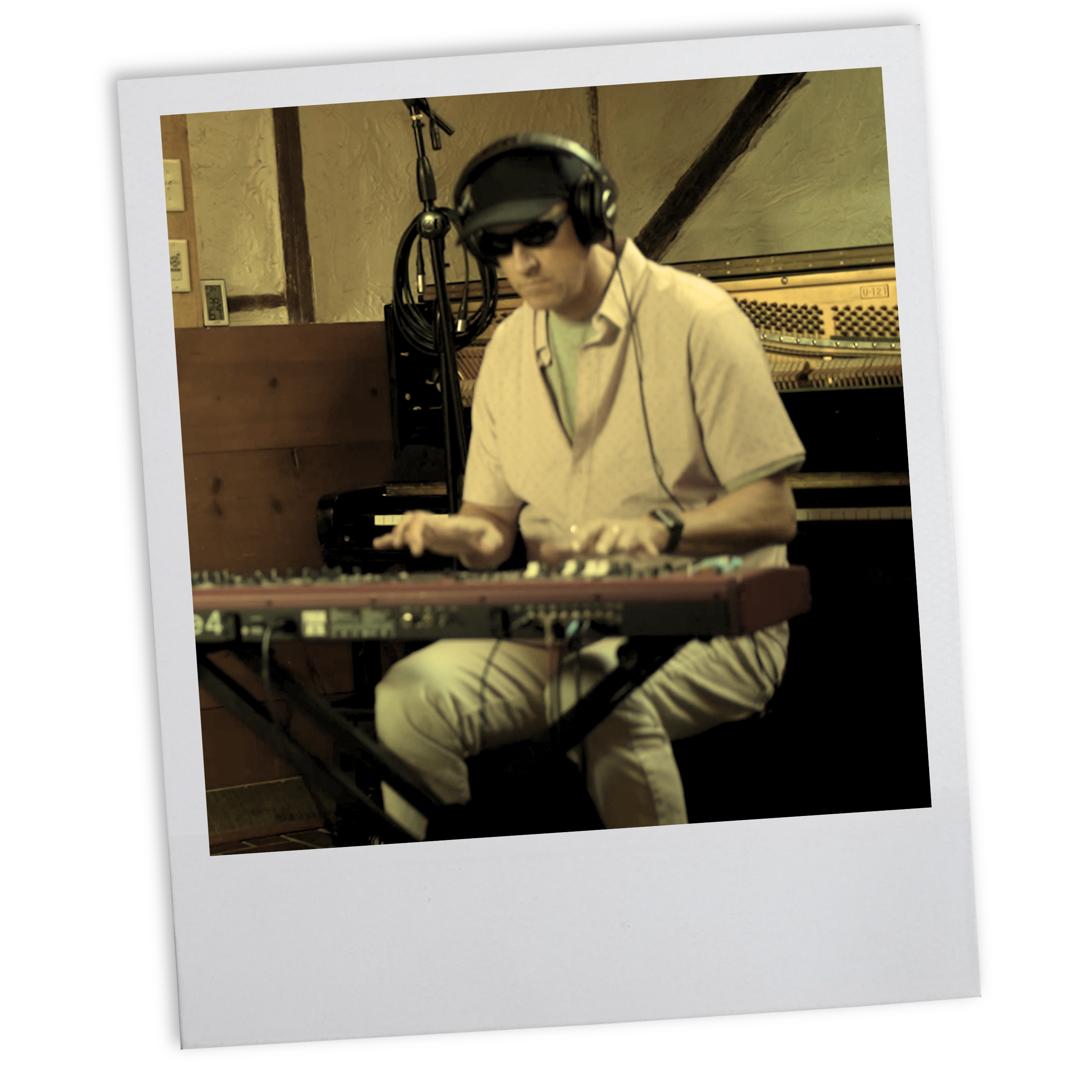A man wearing sunglasses, headphones, and a light-colored shirt playing a keyboard in a recording studio.