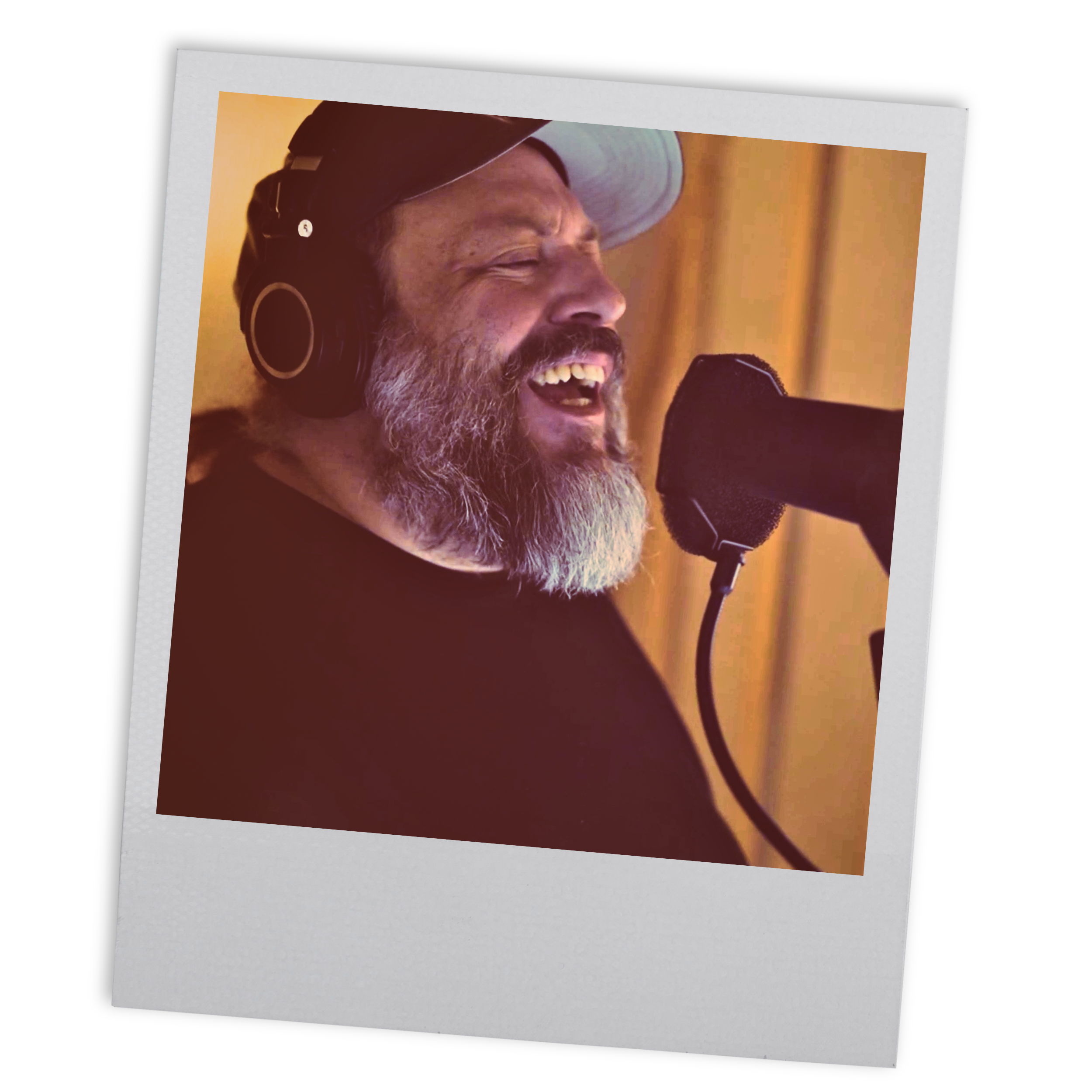 A man with a beard and gray hair singing into a microphone in a recording studio, wearing headphones and a baseball cap.