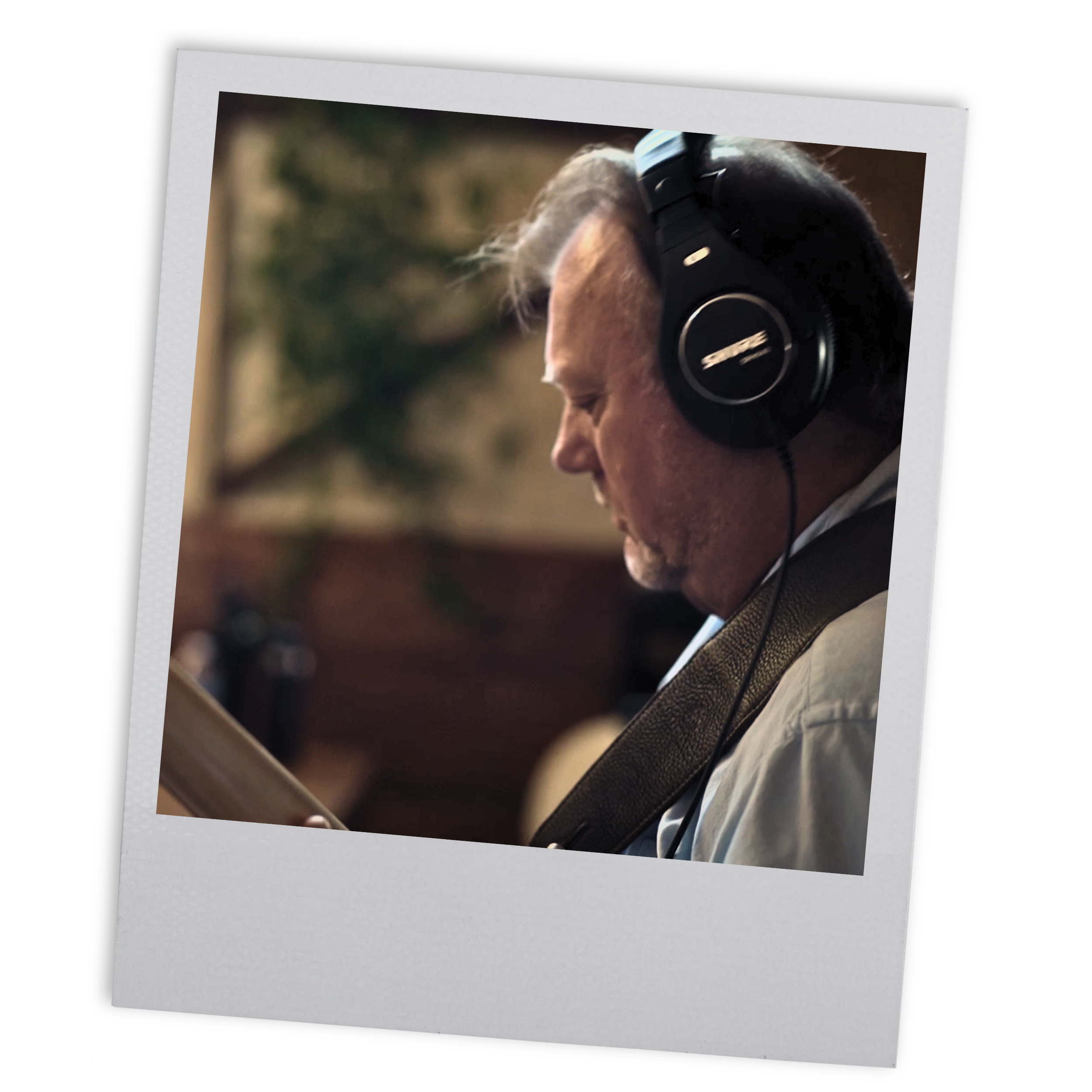 A middle-aged man wearing headphones and a white shirt, reading a book or tablet, seated indoors with a blurred background of a plant and wooden furniture.