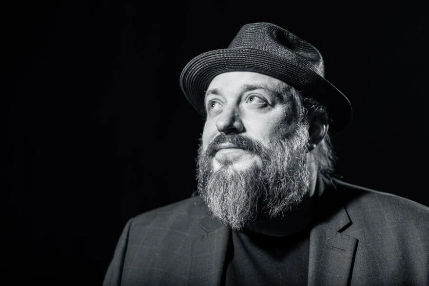 Black and white portrait of a man with a beard and mustache, wearing a fedora hat and a suit, looking to the side with a thoughtful expression.
