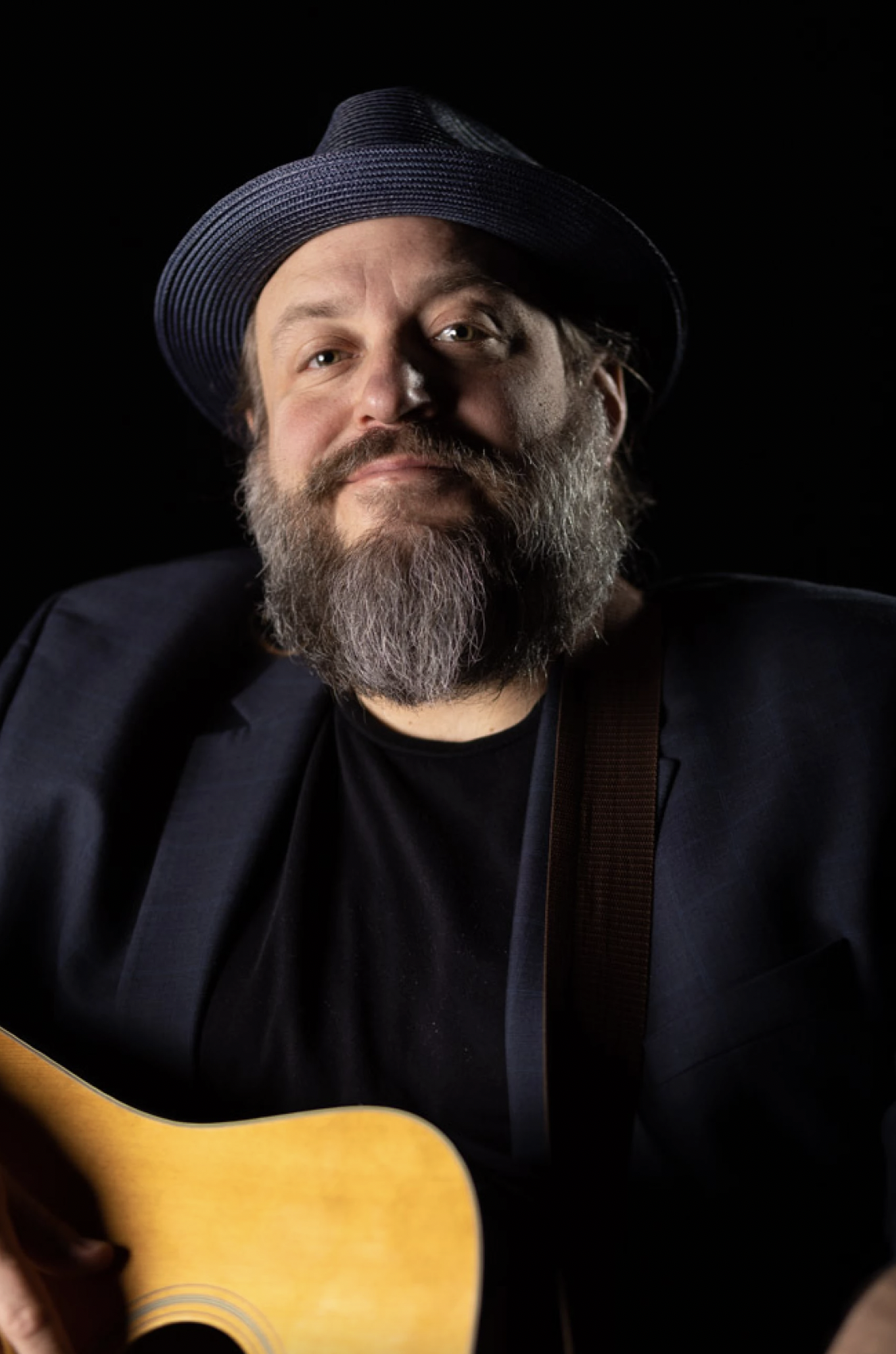 A bearded man wearing a dark hat and black blazer, holding a guitar against a black background.