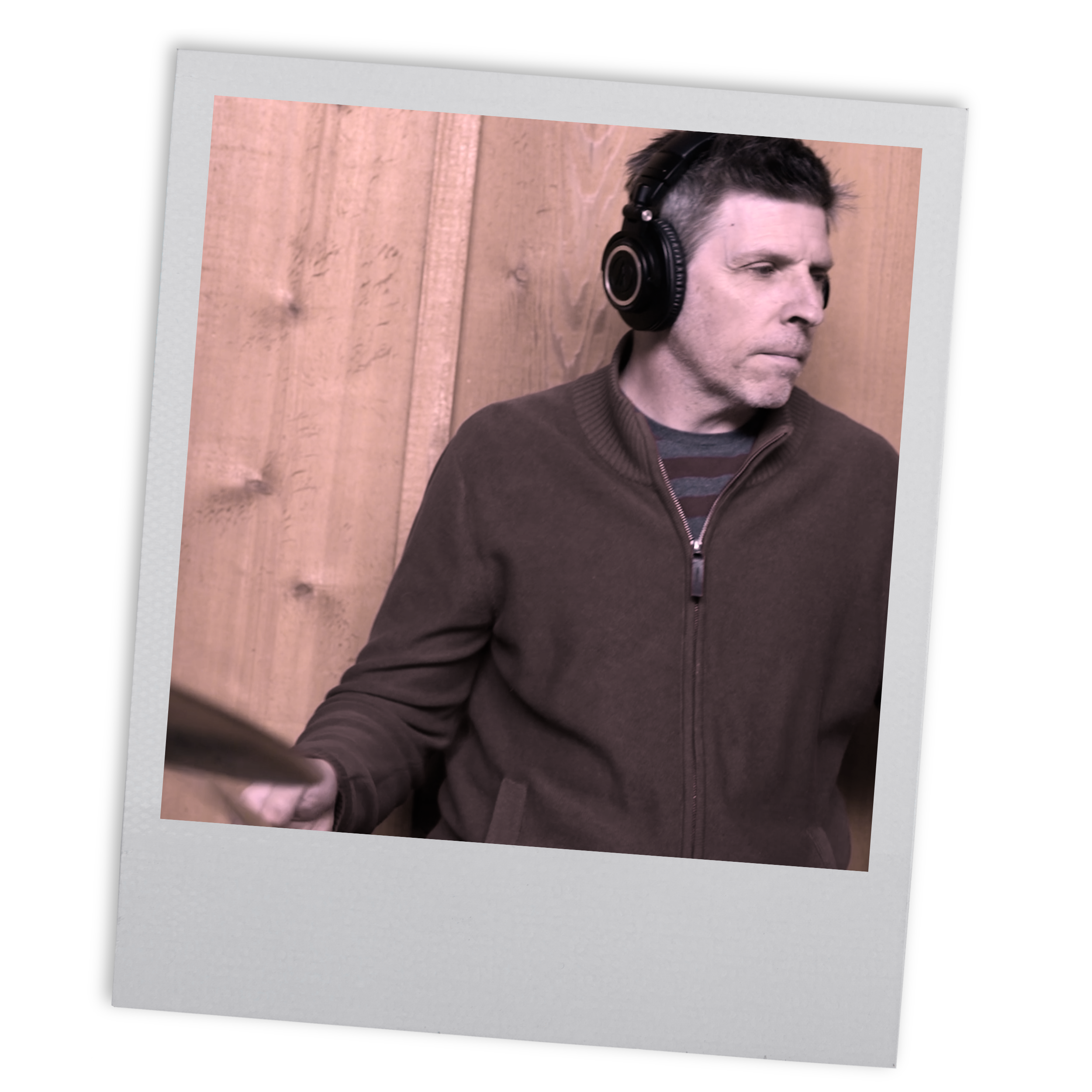 A man with short dark hair wearing a brown zip-up jacket and headphones, holding drumsticks, standing against a wooden wall, looking focused.