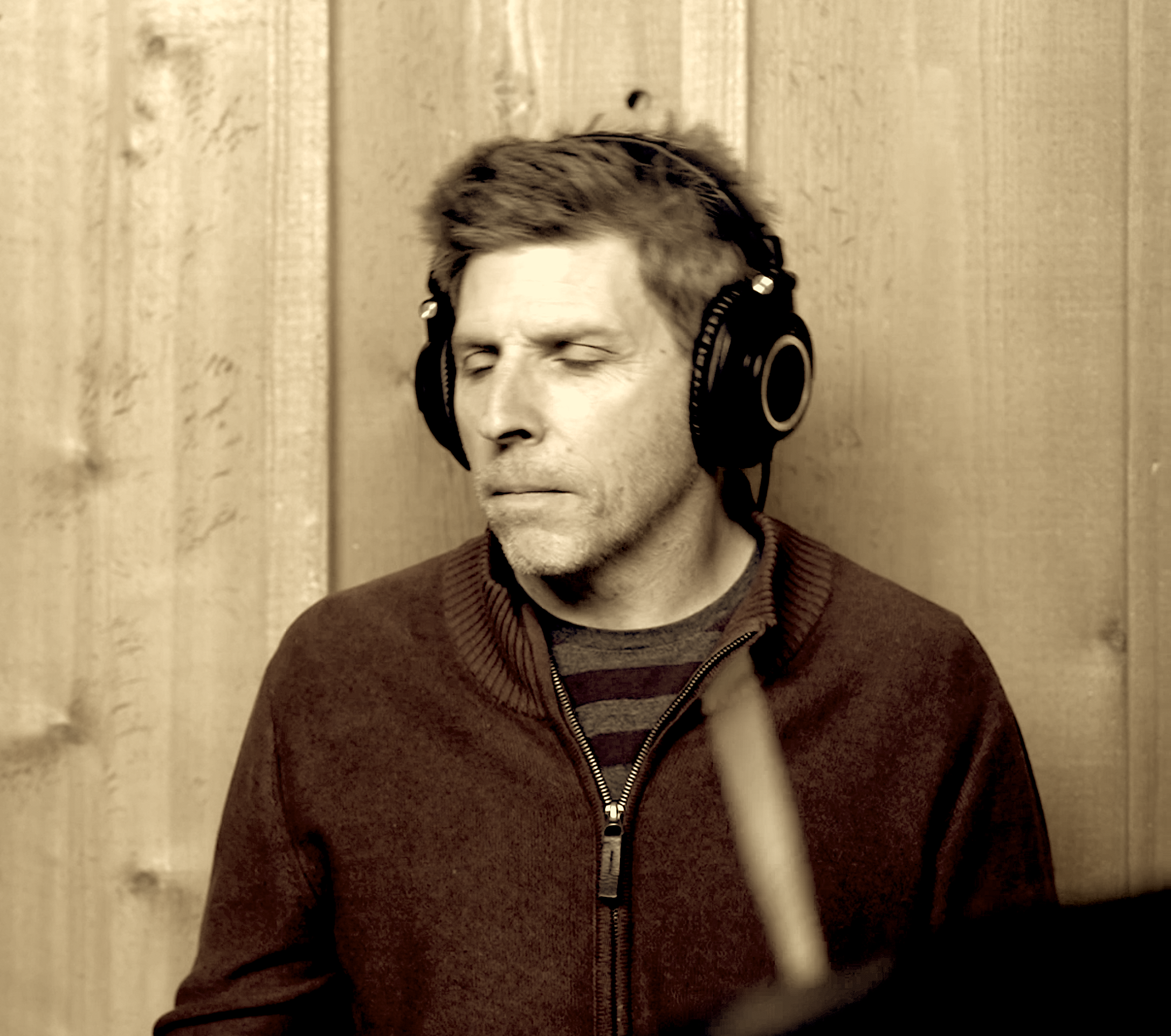 Man wearing headphones with closed eyes, in front of wooden wall, appearing to be enjoying music or sound.