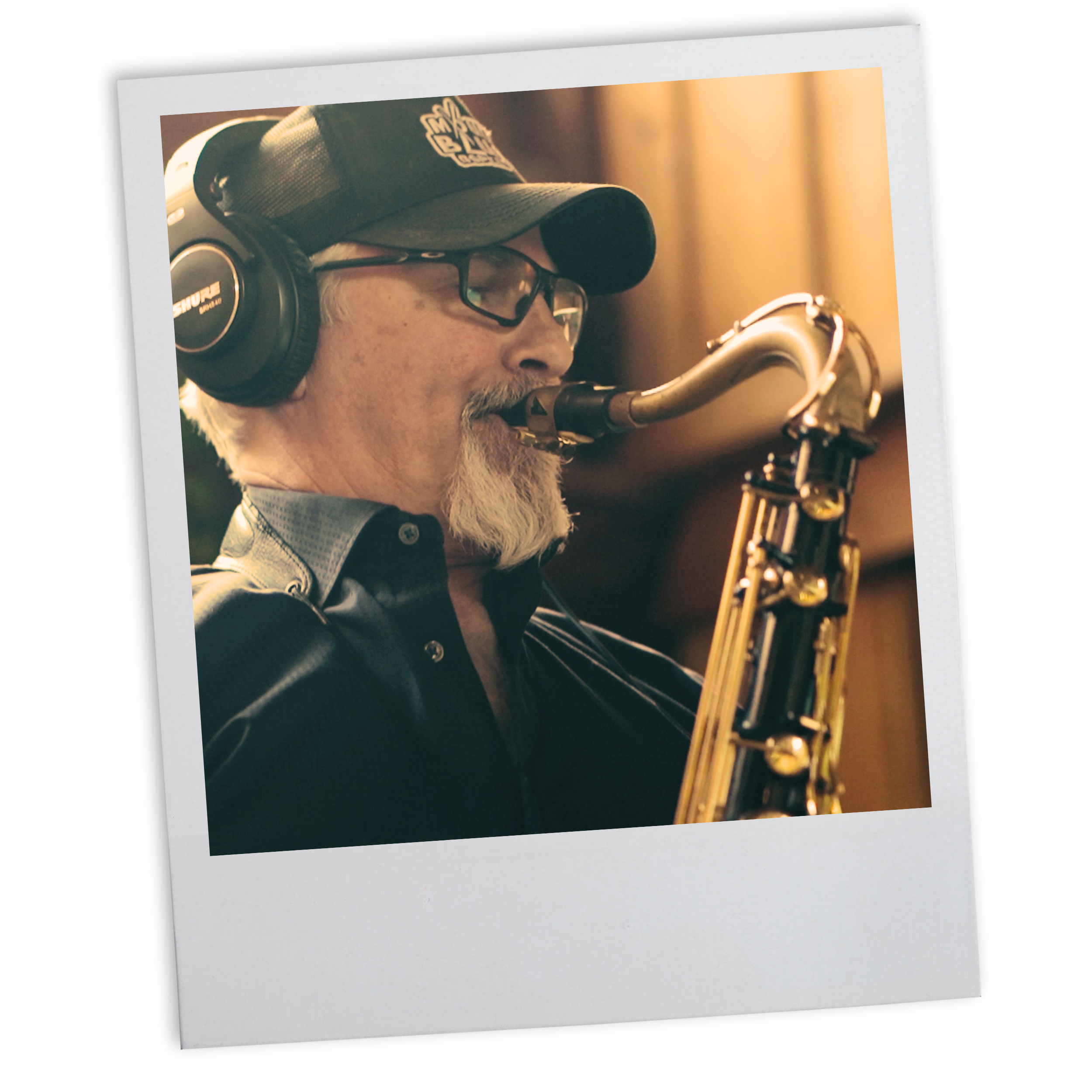 An older man with a gray beard and mustache playing a saxophone, wearing headphones, glasses, a baseball cap, and a leather jacket in a recording studio.