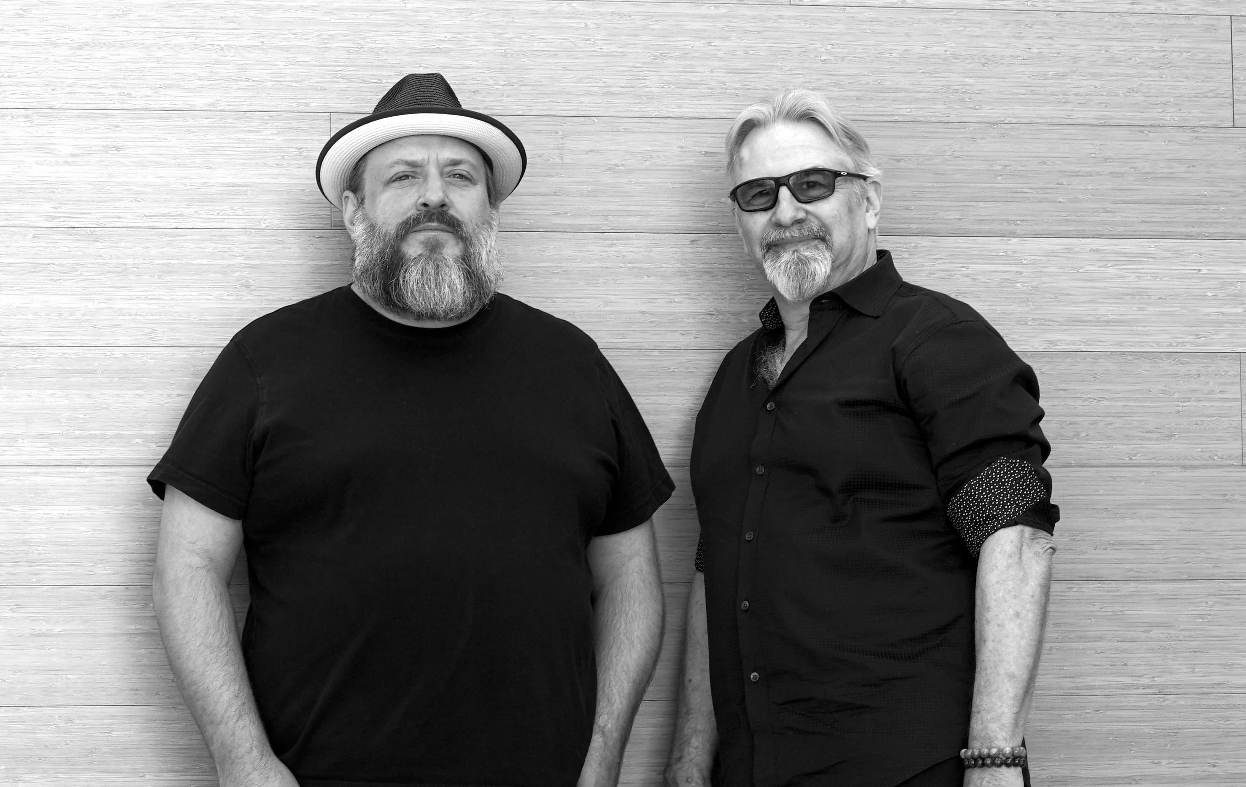 Black and white photo of two men standing against a wood-paneled wall. The man on the left has a beard, mustache, and wears a hat and casual black shirt. The man on the right has glasses, a goatee, and is dressed in a black shirt with rolled-up sleev