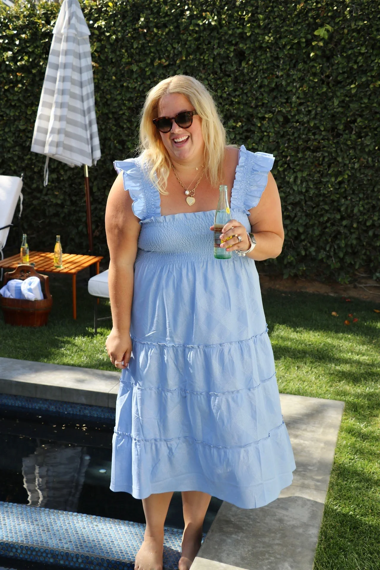 A woman with blonde hair wearing sunglasses and a light blue dress, holding a glass bottle, standing by a poolside outdoors. There are drinks on a small table and a large striped umbrella in the background.