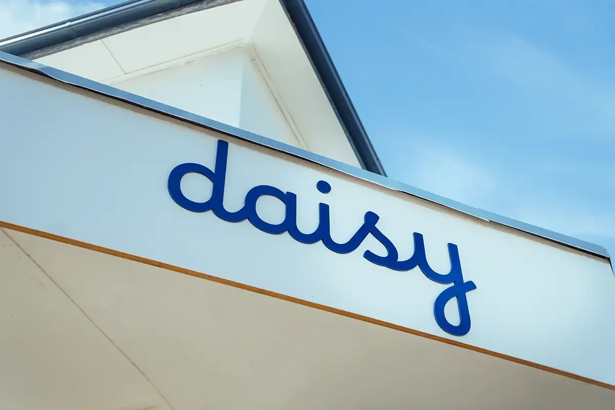 Close-up of a building with a white sign that has the word "daisy" in blue letters, angled with a clear blue sky in the background.