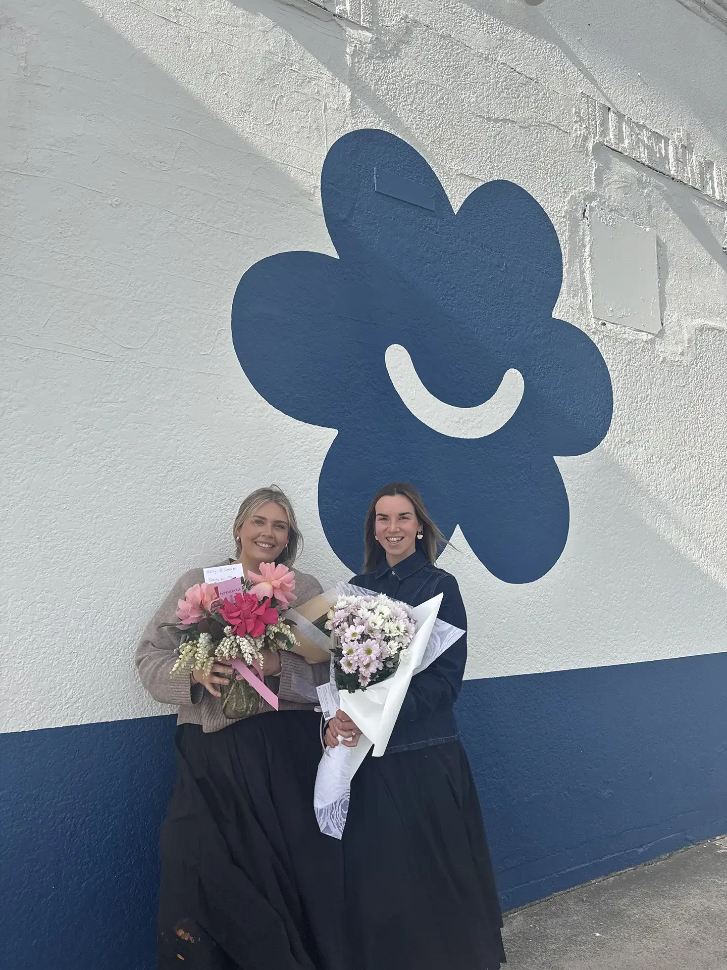 Two women standing outside in front of a wall with a large blue flower logo. They are smiling and each holding a bouquet of flowers. One woman is wearing a beige sweater and a black skirt, the other is wearing a dark shirt and a long dark skirt.