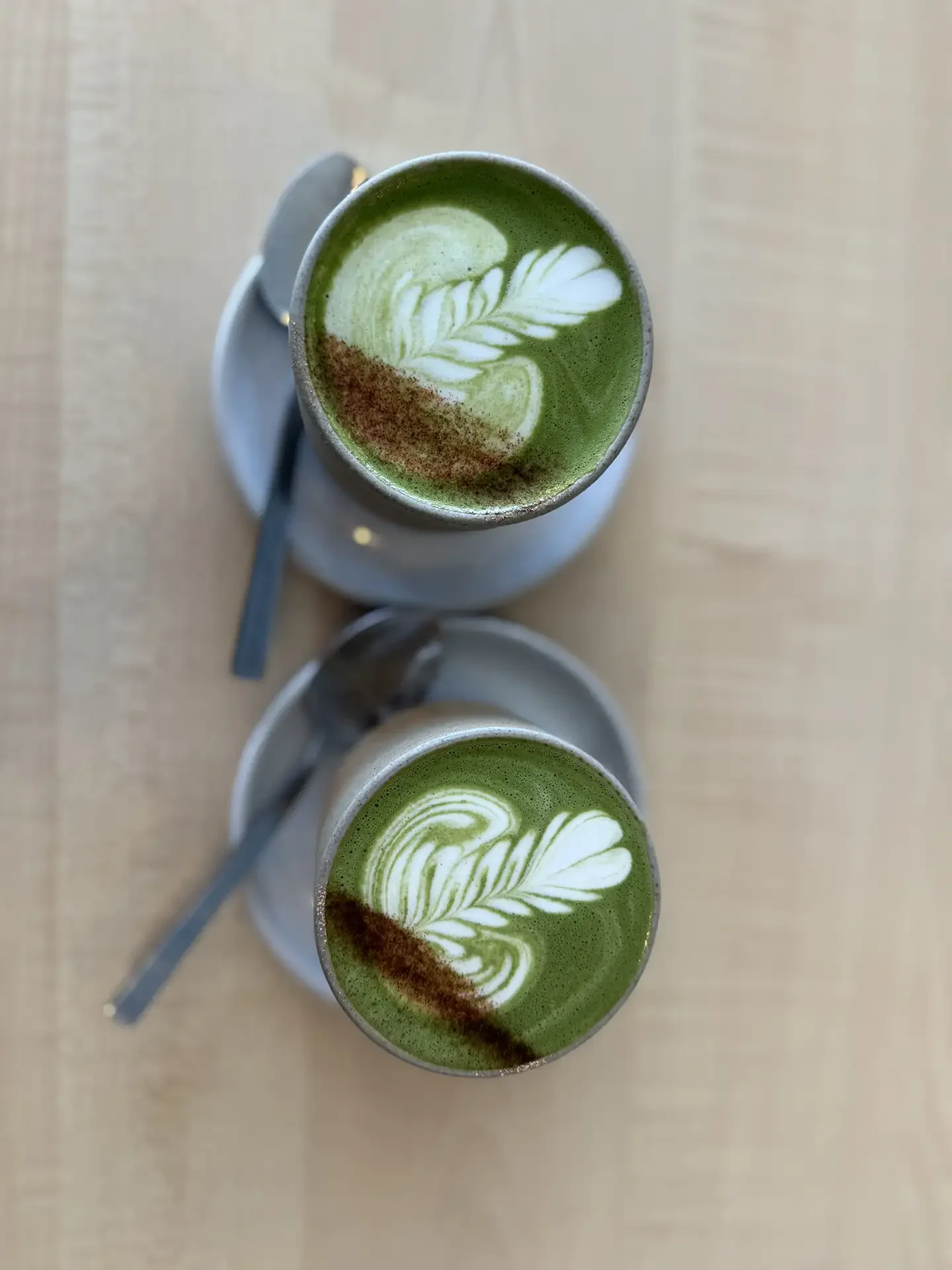 Two cups of matcha latte with latte art on top, placed on a wooden surface. Each cup has a spoon next to it.