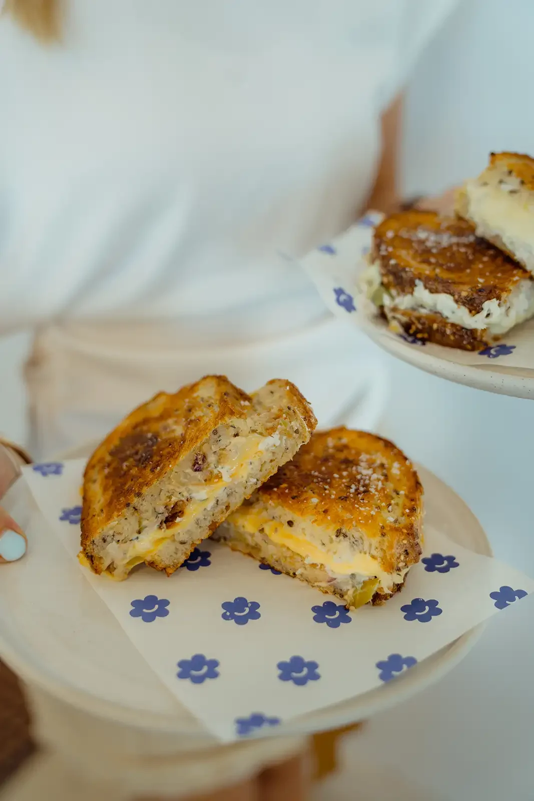 Close-up of a plate with a breakfast sandwich filled with eggs, cheese, and sausage, with a second sandwich split in half showing the filling, on decorative paper with blue flowers.