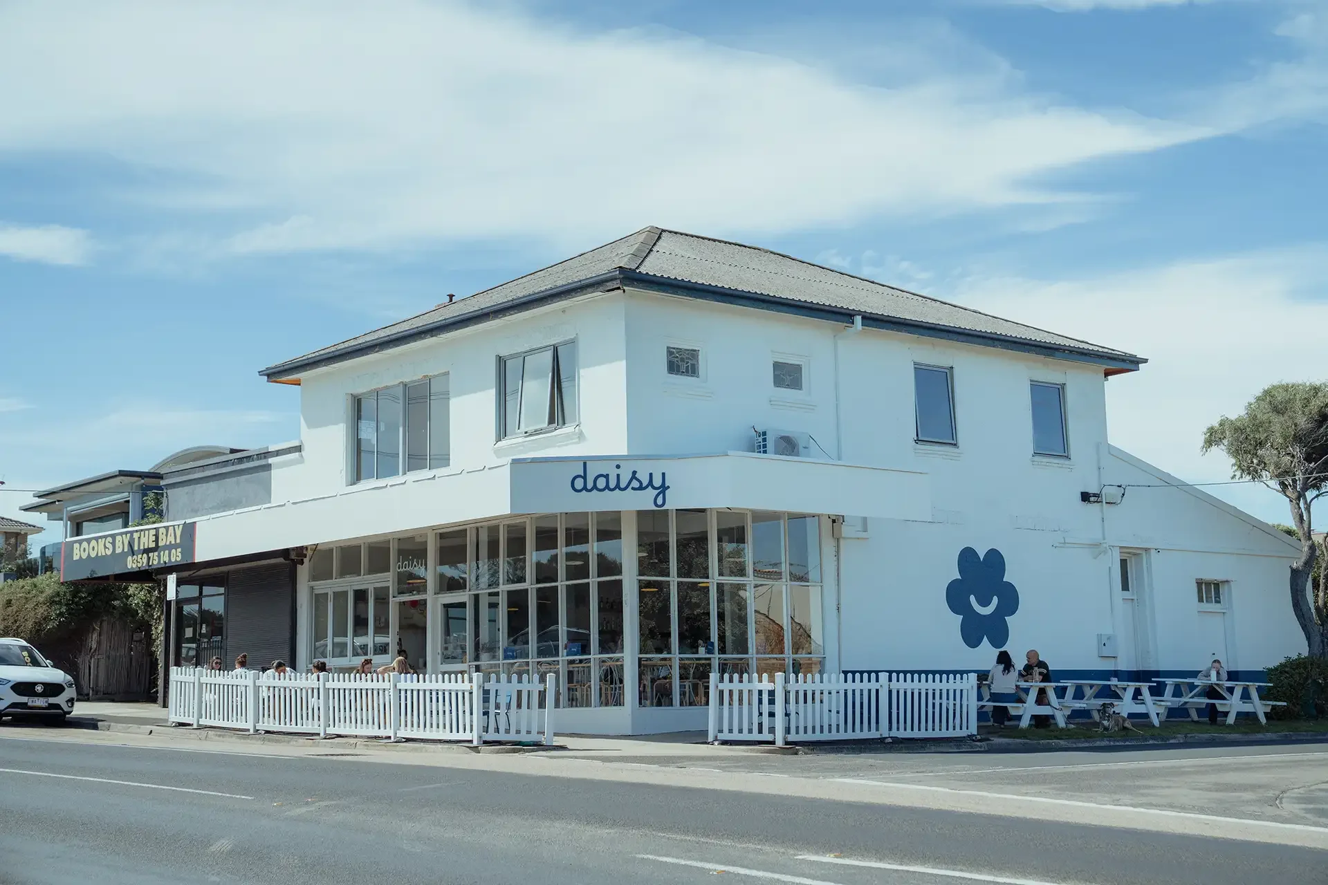 The exterior of a two-story building with a white facade, large glass windows on the ground floor, and the name ''daisy'' written on it. There is a blue flower logo on the wall, a white picket fence surrounding an outdoor seating area with tables and people, and a sign for a bookstore called ''Books by the Bay.'' The sky is partly cloudy with blue and white hues.