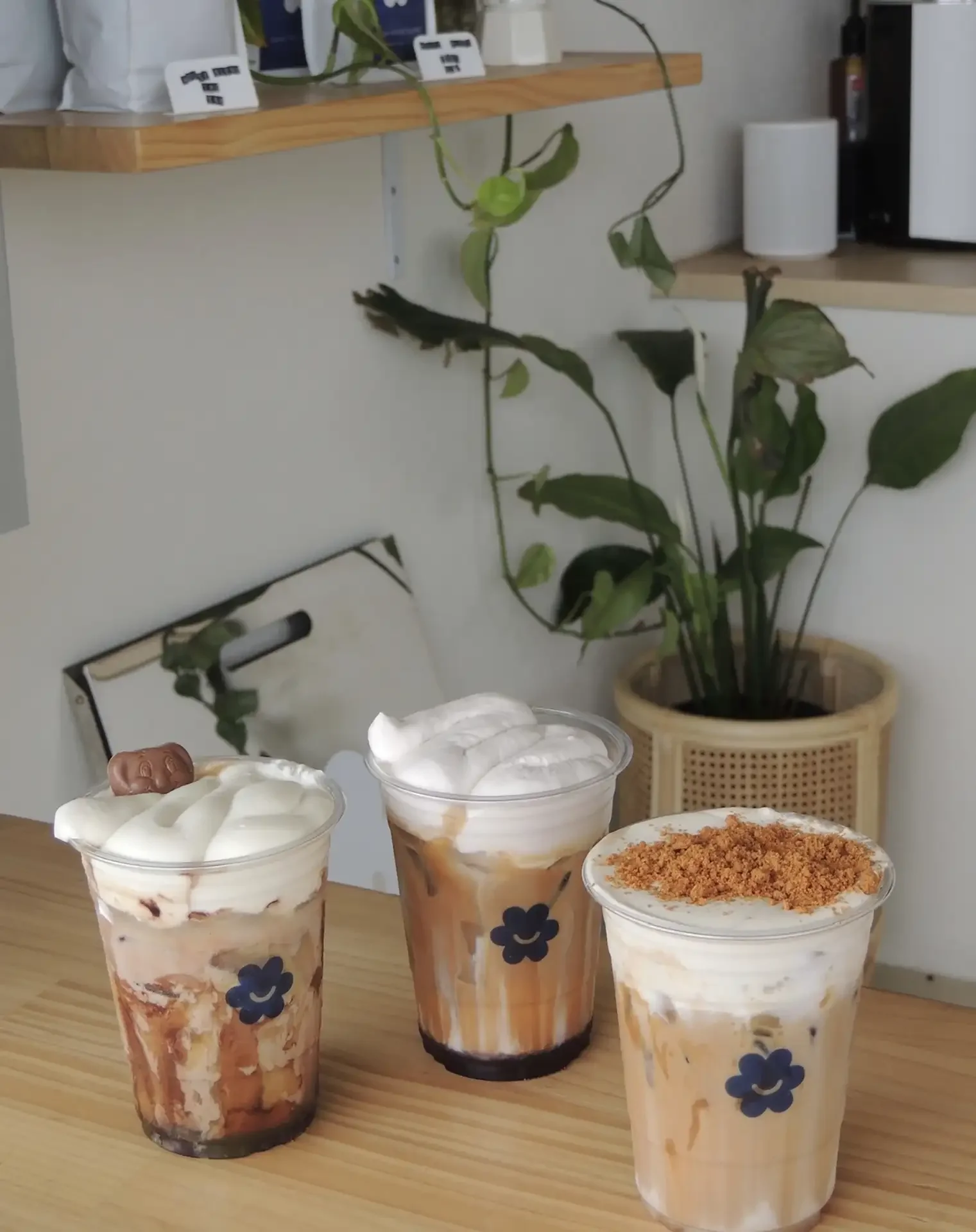 Four milkshakes with whipped cream and toppings on a wooden table, with a framed mirror, plant, and shelves in the background.