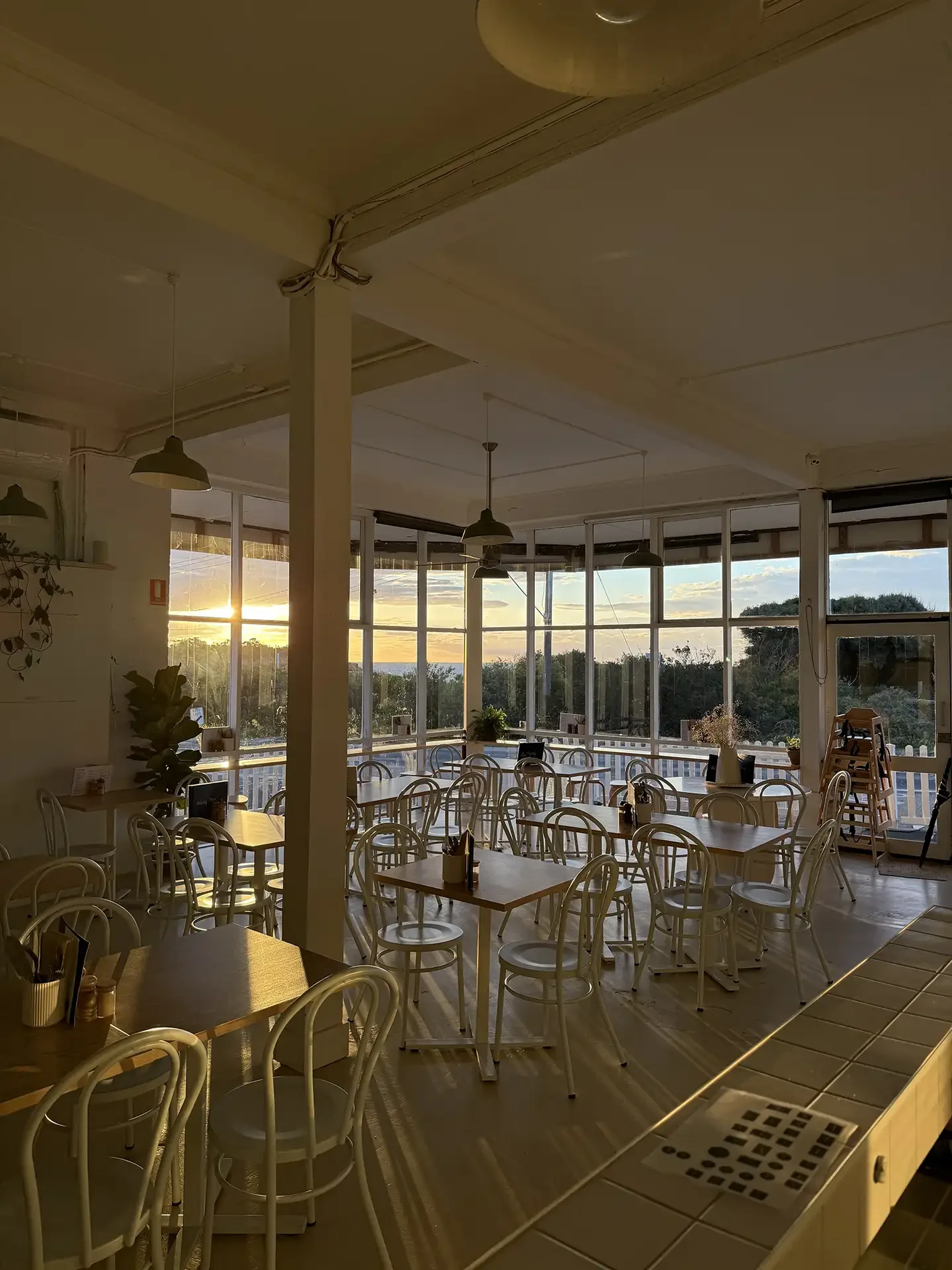 Interior of a cozy cafe with white chairs and wooden tables, large windows showing a sunset over greenery, and warm lighting.