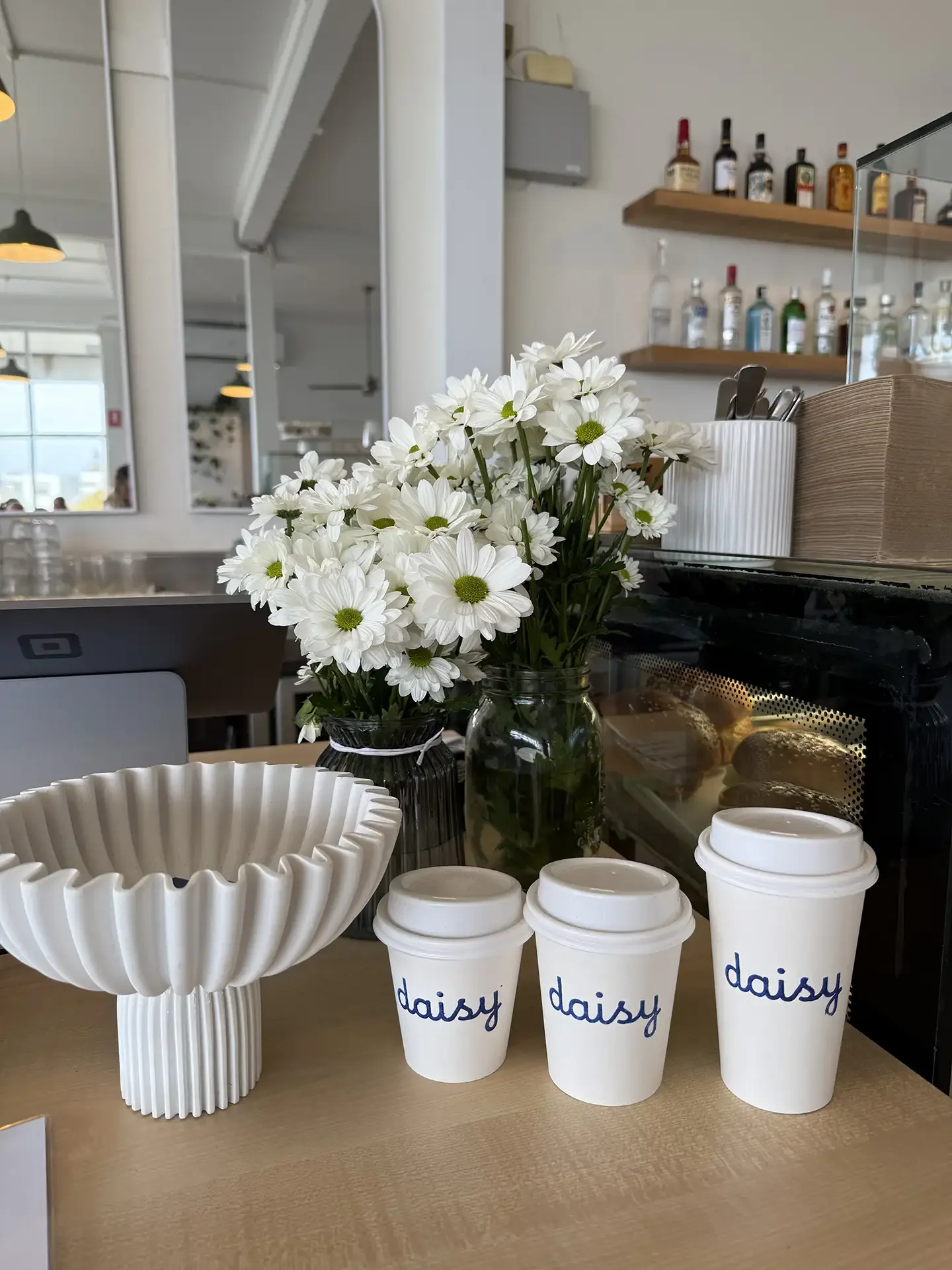 A vase of white daisies, three white disposable coffee cups with 'daisy' written on them, and a fluted white ceramic bowl on a light wood table inside a cafe.