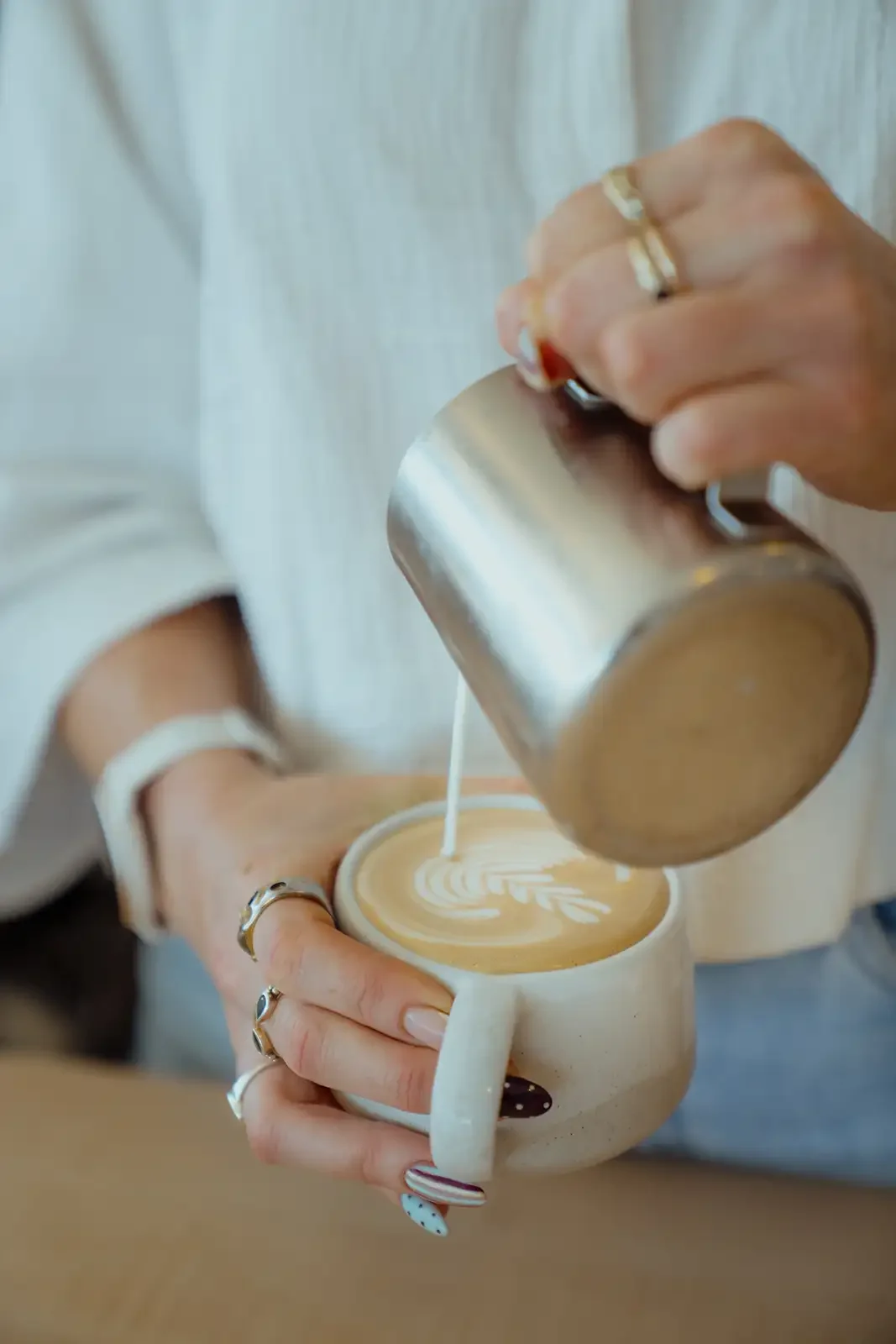 A person pouring steamed milk into a latte, creating latte art in a beige mug, with the person wearing rings on fingers and a white shirt.