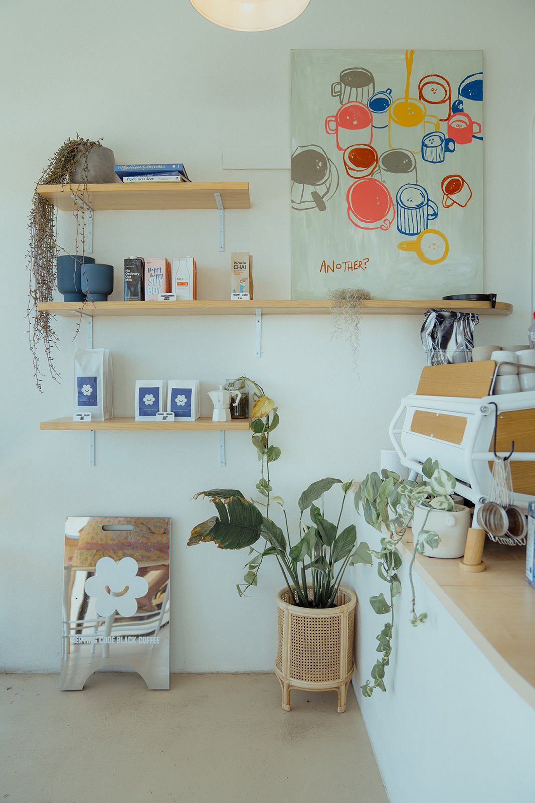Interior of a cozy coffee shop with wooden shelves holding cups, plants and coffee supplies, a colorful wall art featuring coffee mugs, and a plant in a decorative planter near the counter.