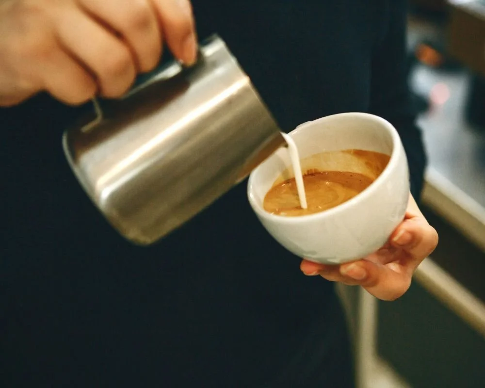 A person pouring steamed milk into a cup of coffee. 