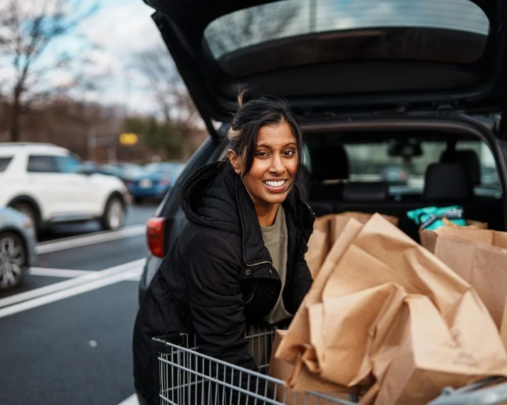 A woman loading grocery bags into an open trunk of a car at a parking lot.