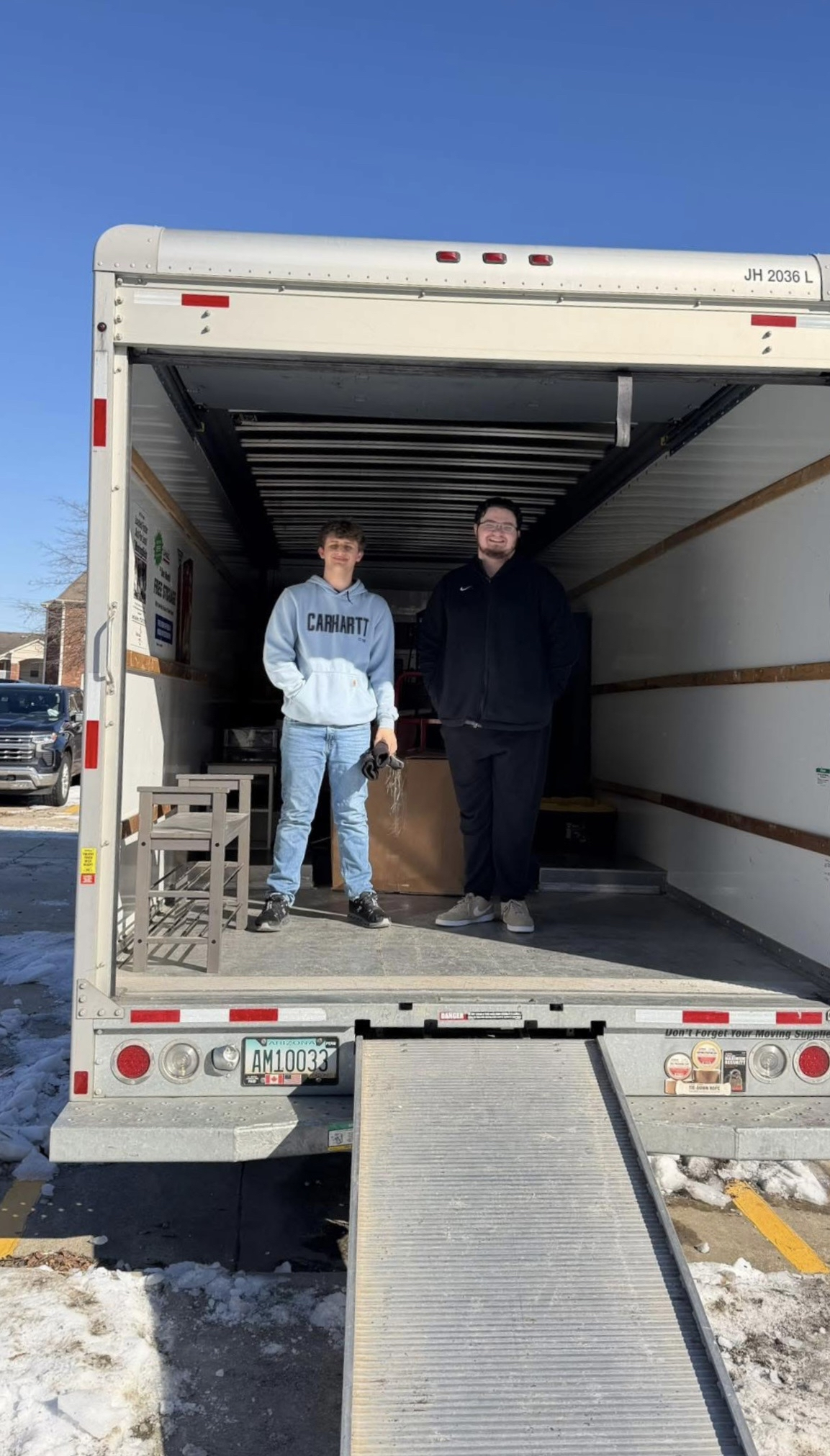 Two people are standing inside the open back of a moving truck parked outside in a parking lot, with one person holding car keys and the other smiling. There is snow on the ground nearby.