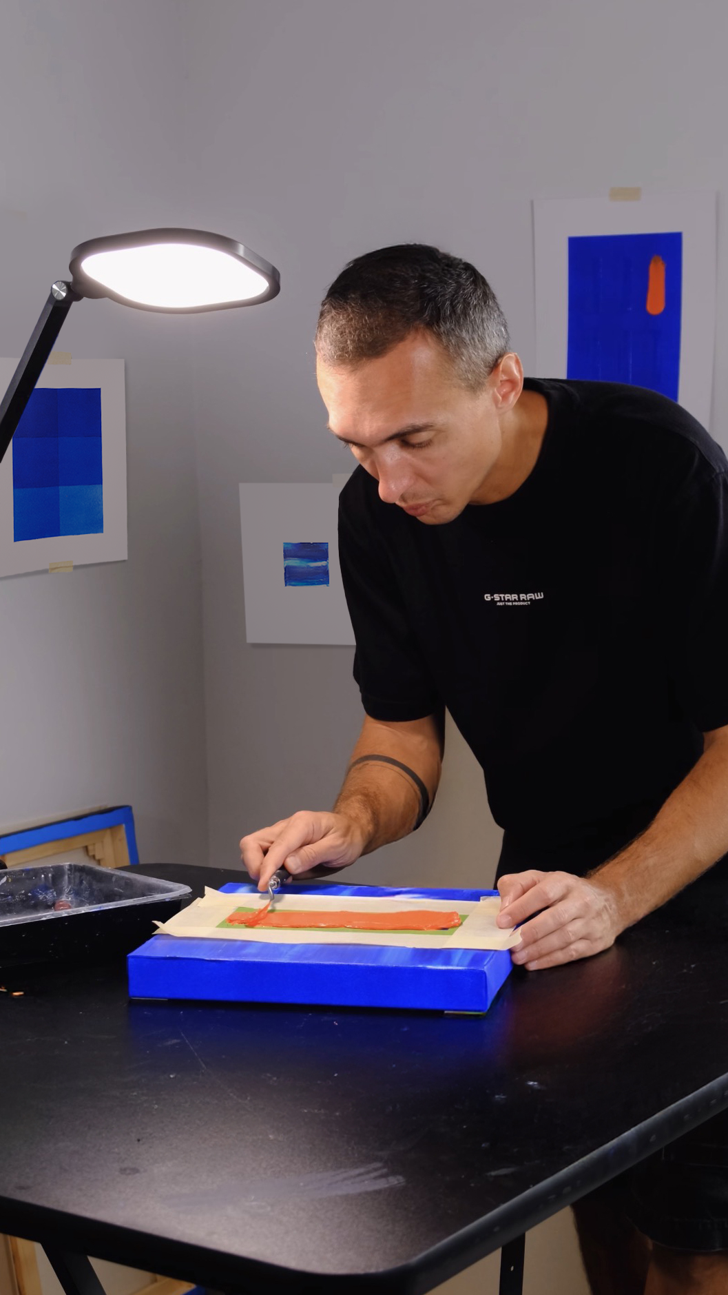 A man is creating a piece of artwork using a tool on a piece of paper or canvas on a table, with blue and green colors visible. The room has artwork displayed on the walls and a bright light overhead.