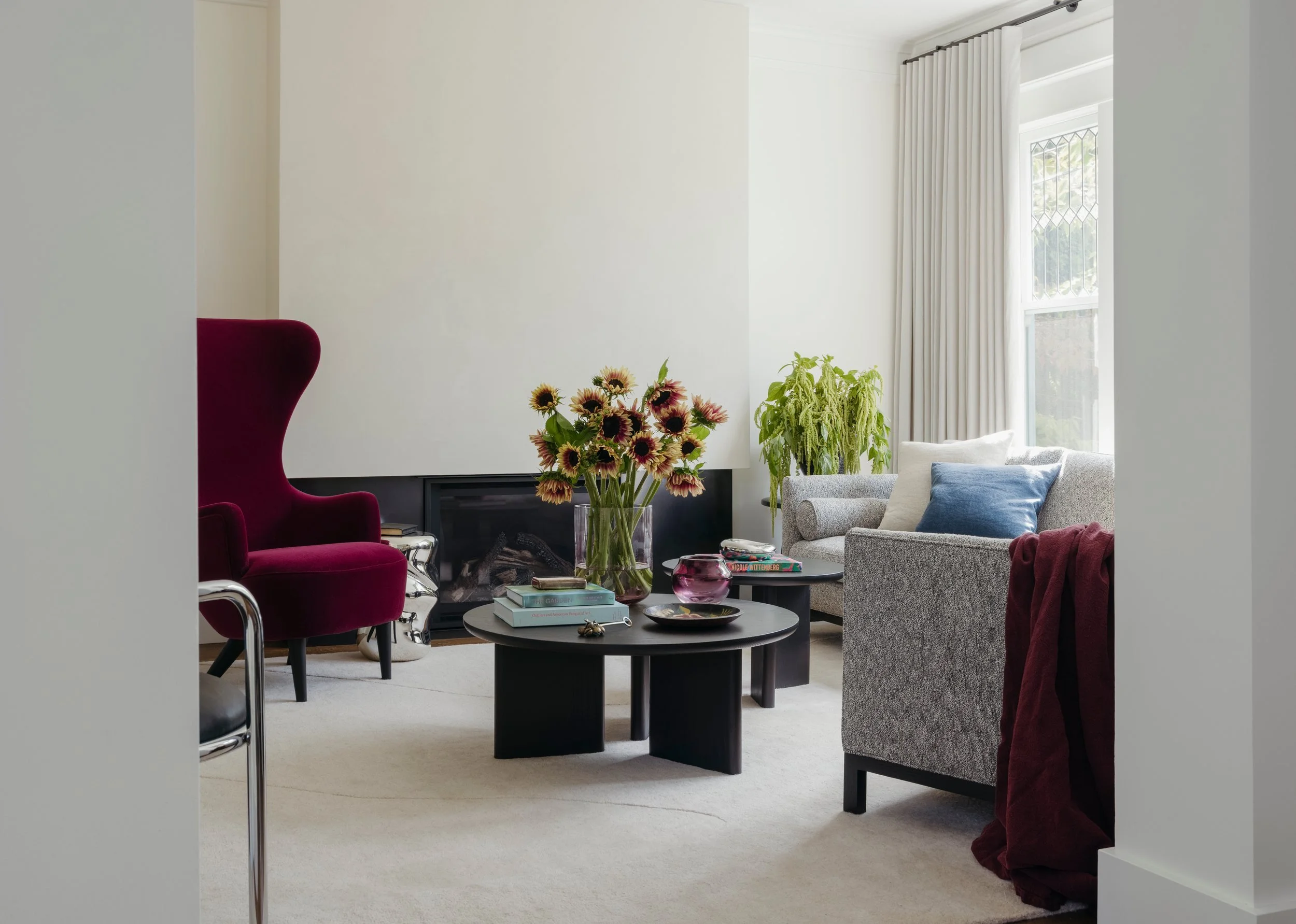 View into the living room past a burgundy wing chair, with the fireplace, round coffee tables and taupe drapery