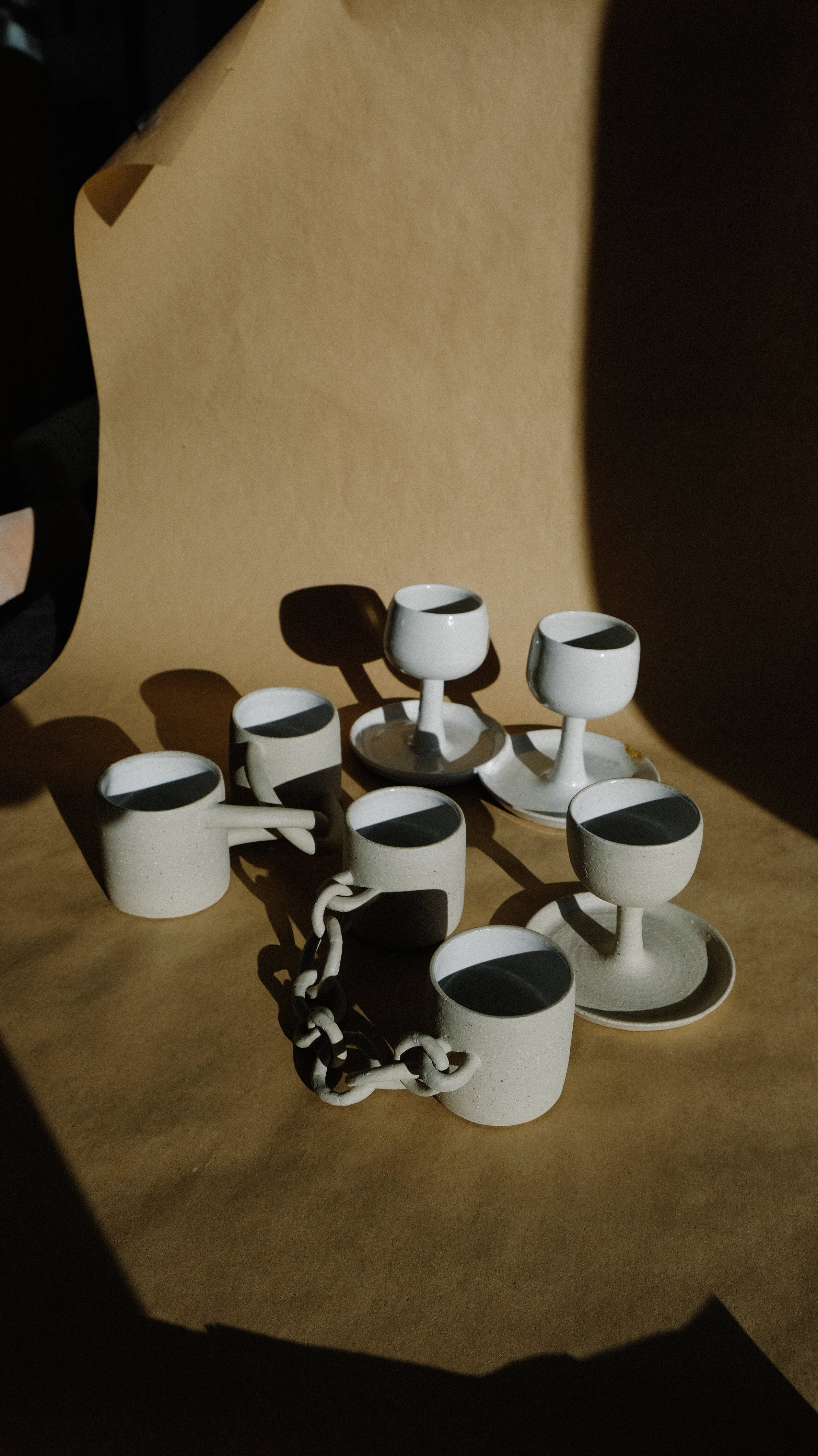 White ceramic cups and small plates with dark liquid, arranged on a brown surface with dramatic lighting creating shadows.