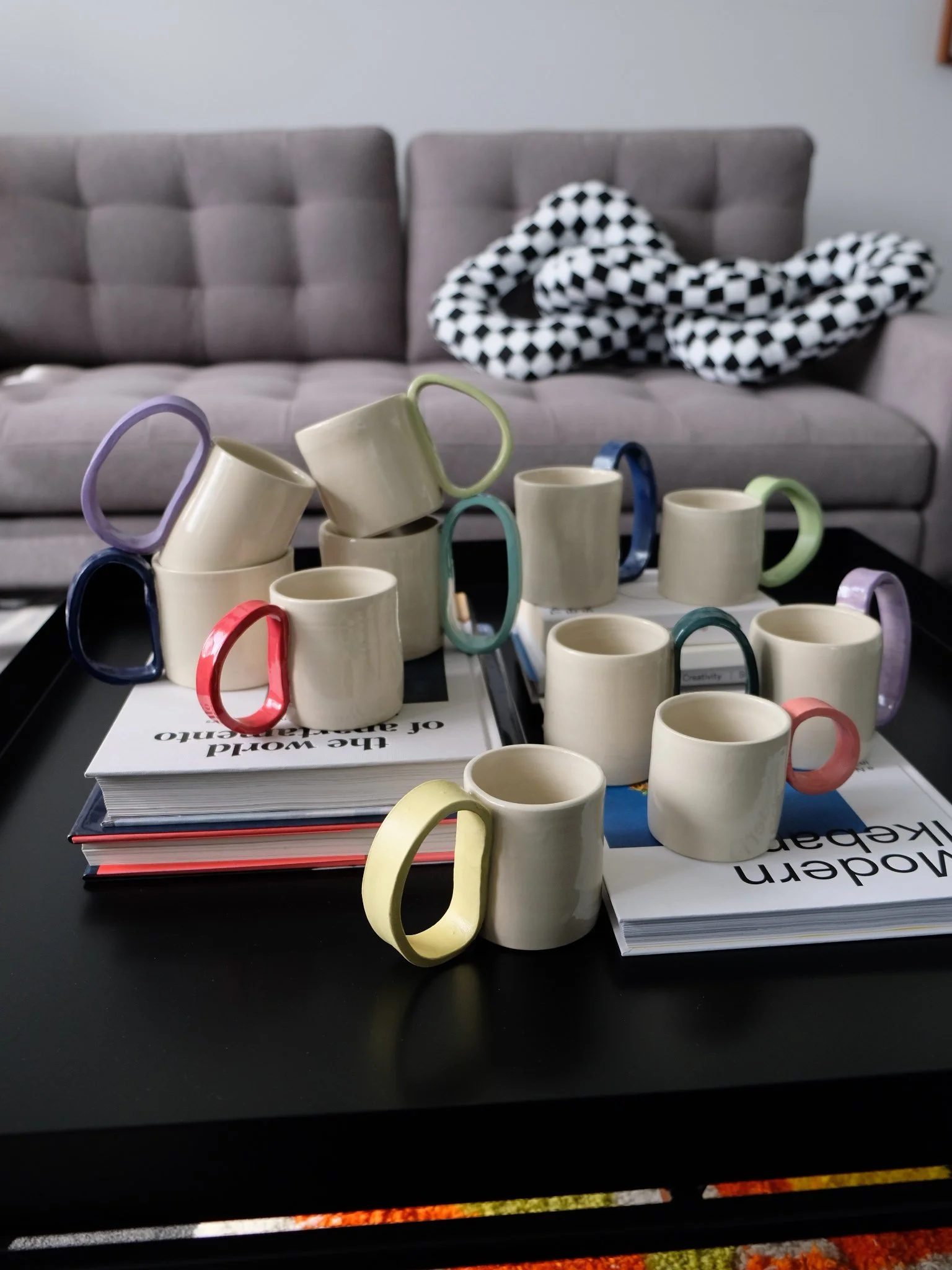 Multiple ceramic mugs with colorful handles placed on top of stacked magazines on a black coffee table in a living room, with a gray sofa and a black-and-white checkerboard blanket in the background.