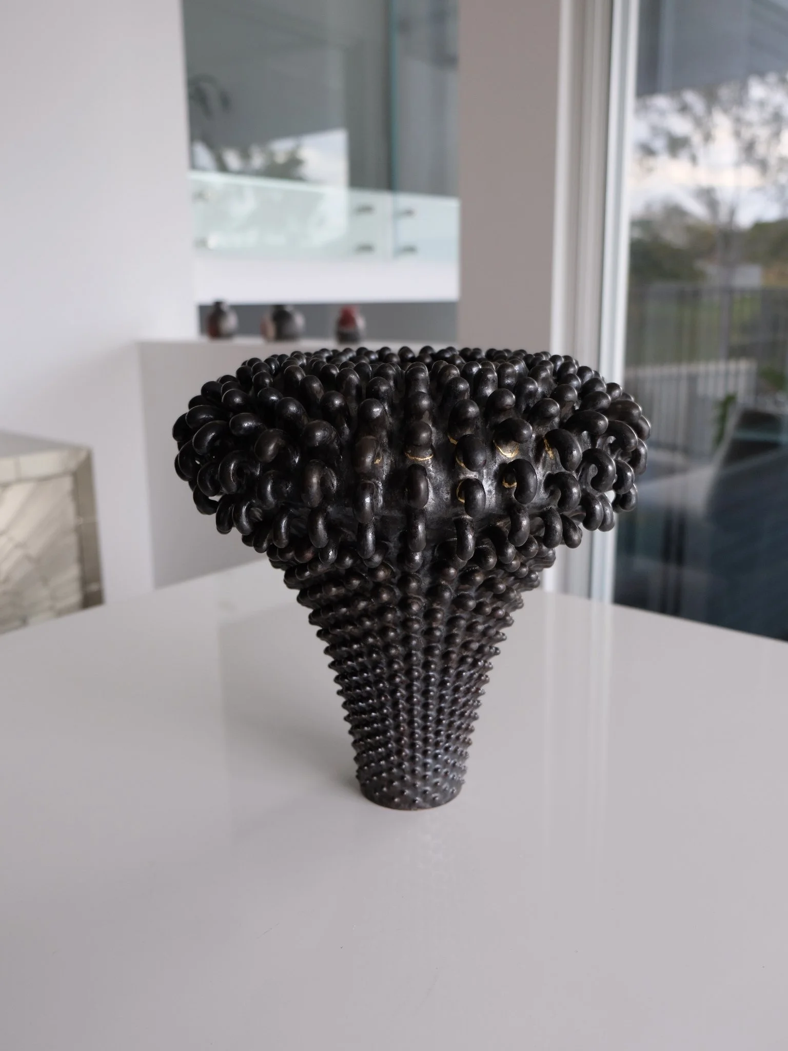 Black textured vase with raised circular patterns, placed on a white surface in a modern indoor space with large windows.