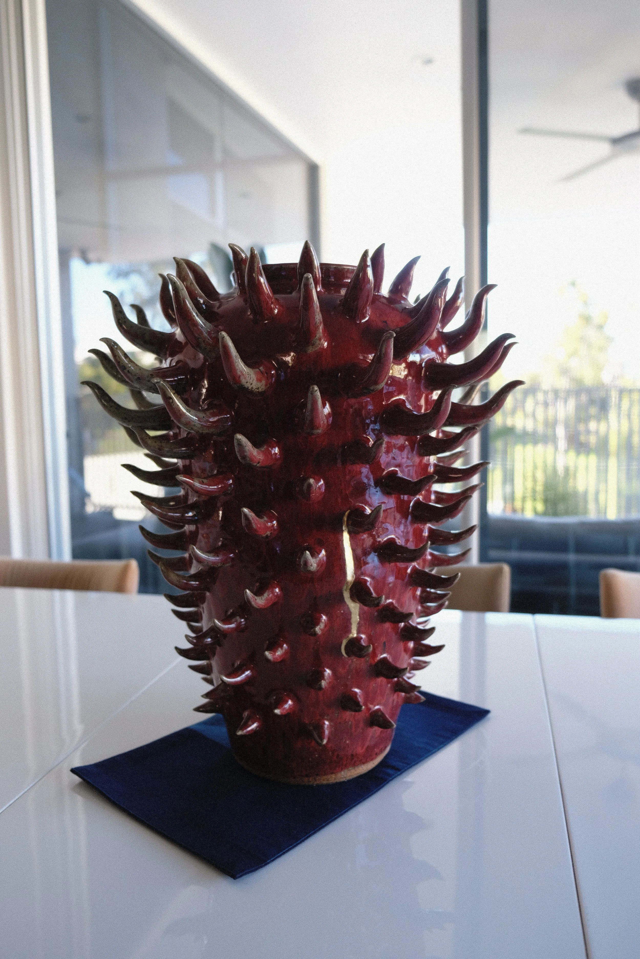 A large, red vase with numerous dark, curved protrusions on a white table with a dark cloth underneath, in a room with large windows and an outdoor view. Kintsugi decorative ceramic repair, gold line repair.