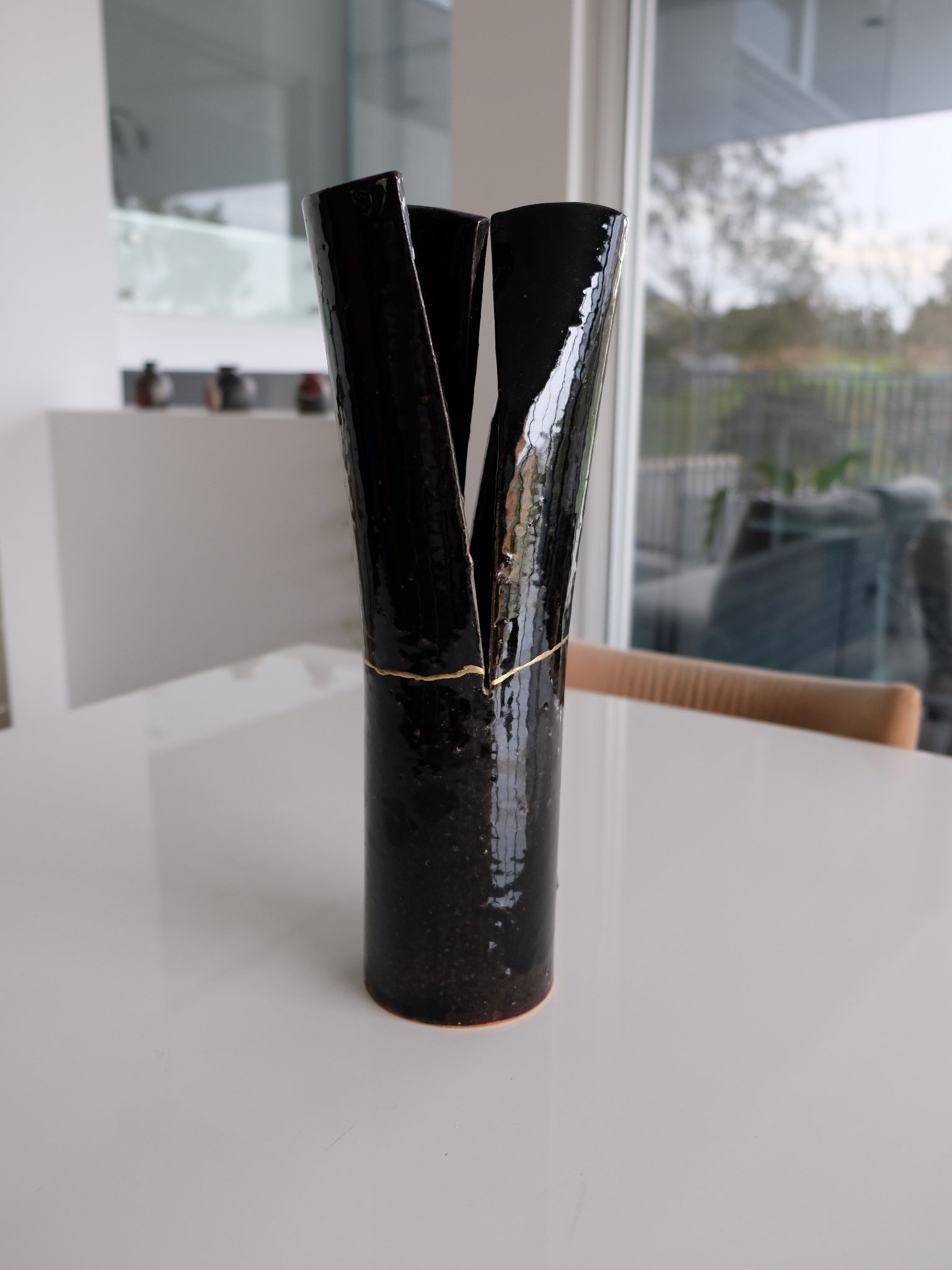 A black ceramic vase with three tall, curved, and shiny surface panels, placed on a white table in a modern interior with large windows.