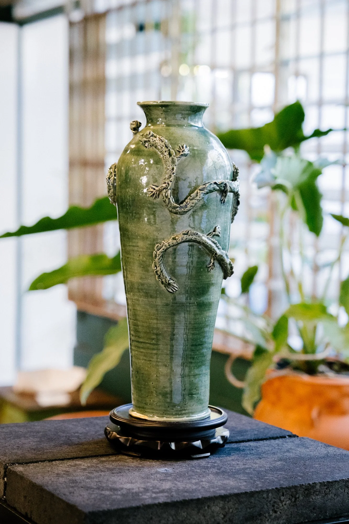 A tall, green ceramic vase with decorative lizard sculptures attached to its surface, placed on a black display stand on a dark table, with plants and a wooden lattice background.