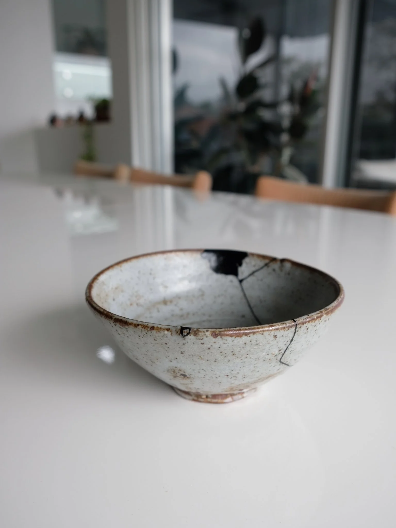 A ceramic bowl with visible cracks and a weathered, rustic appearance, placed on a white surface with a blurred modern indoor background. Black kintusgi ceramic repair, with urushi, food safe.