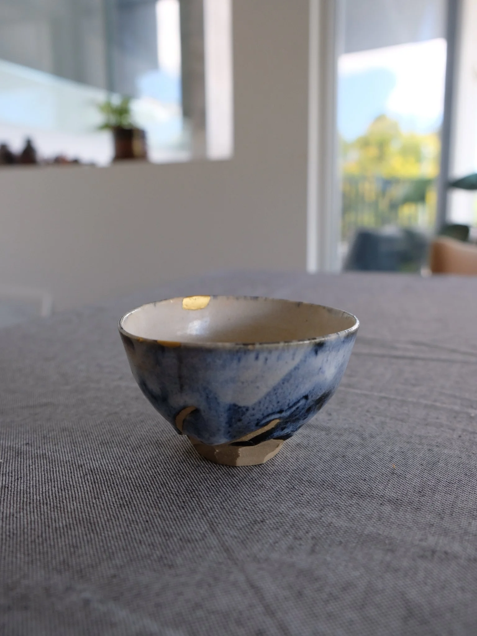 A small ceramic bowl with a blue and white marbled pattern placed on a gray tablecloth in a sunlit room with large windows and a potted plant in the background.