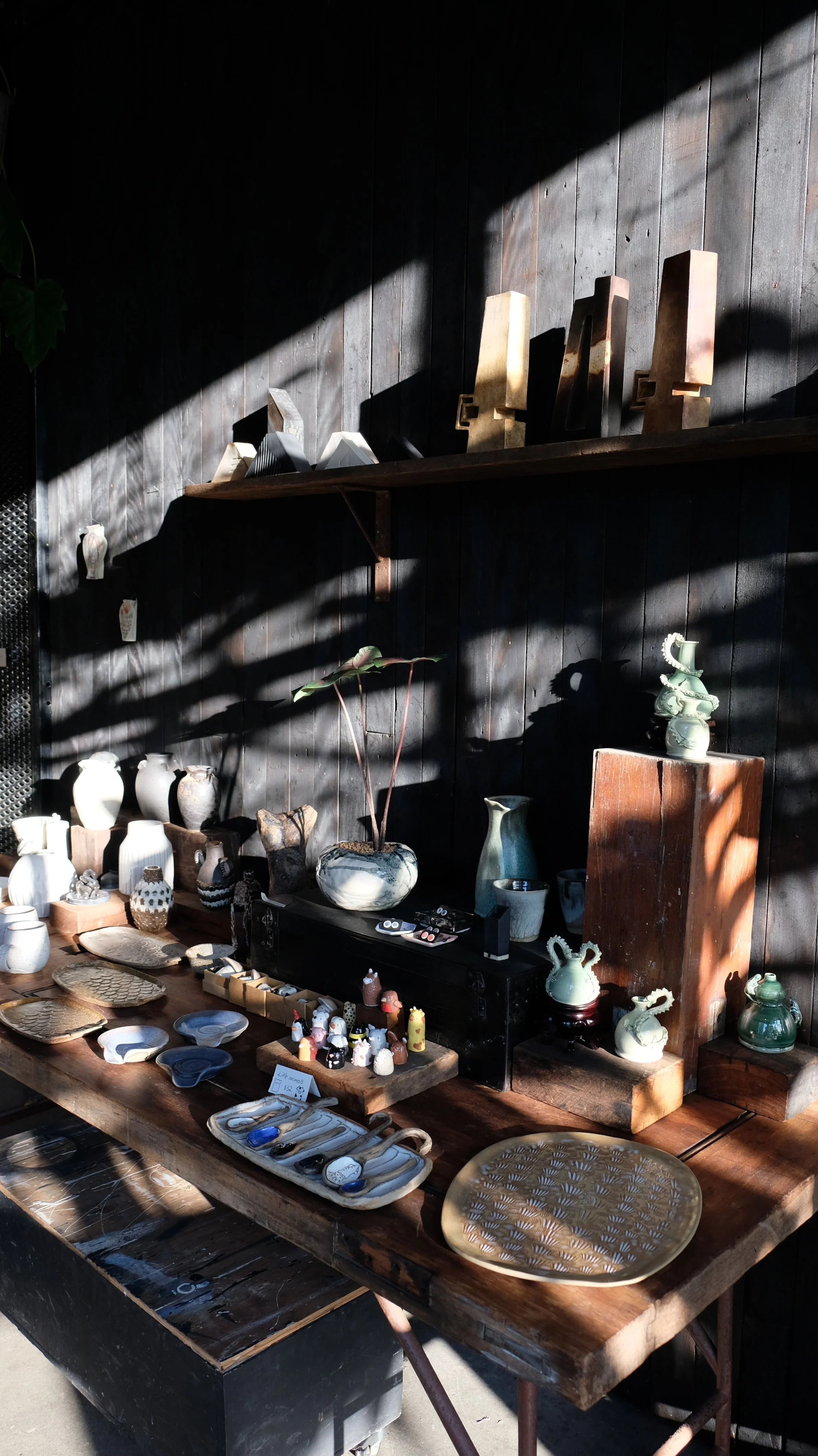 A wooden table displaying various ceramics and decorative items, with a dark wooden wall and shelving behind it, partially illuminated by sunlight.