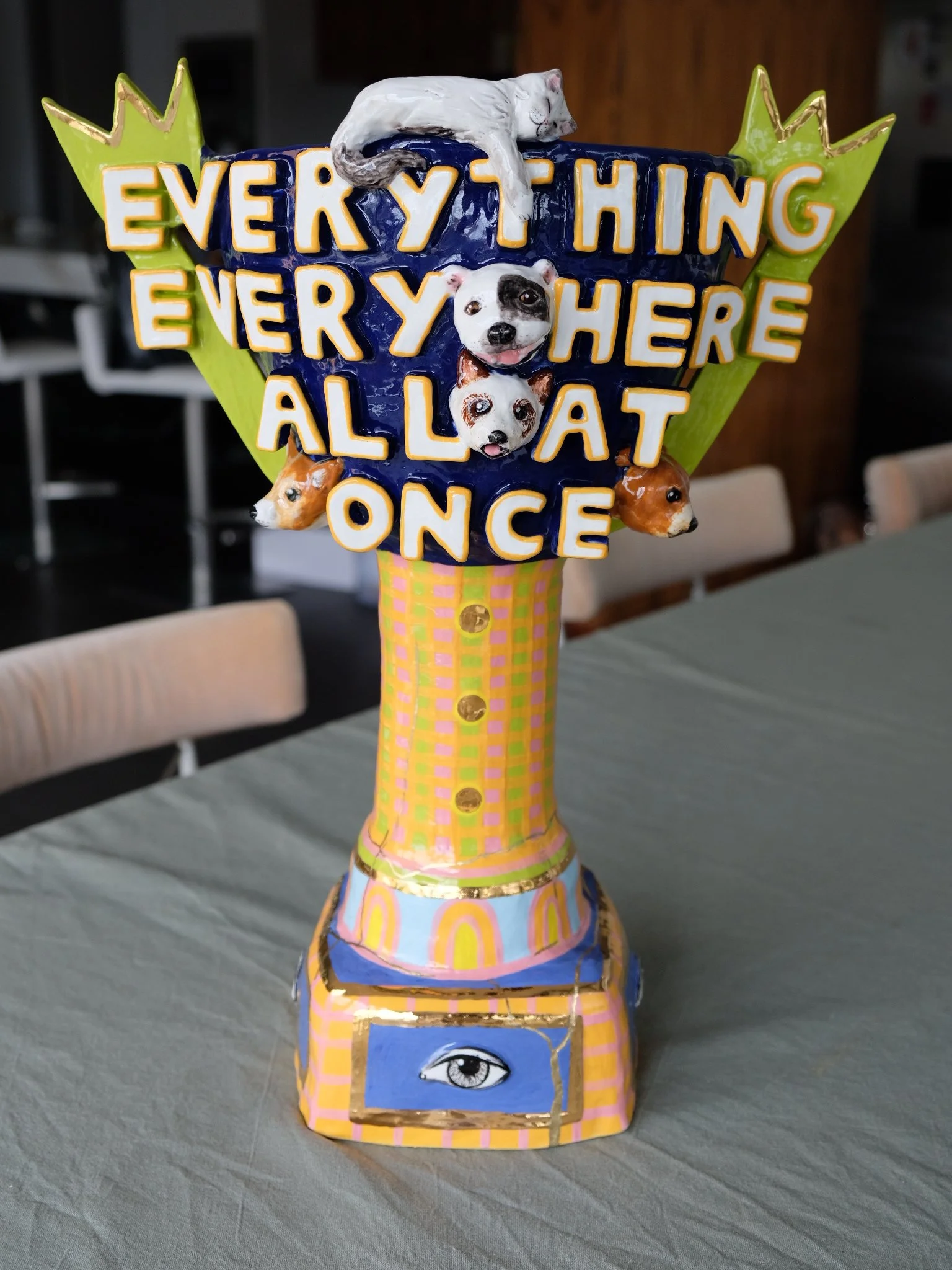 Colorful decorative sculpture with a sign that reads "Everything everywhere all at once", featuring dog faces and an eye symbol at the base, displayed on a table in a room.