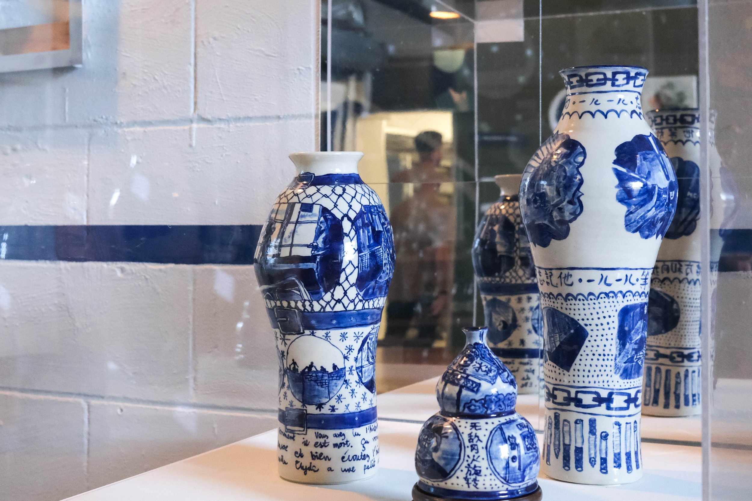 Collection of blue and white decorative ceramic vases displayed in a glass case, with reflections and a blurred person visible in the background.