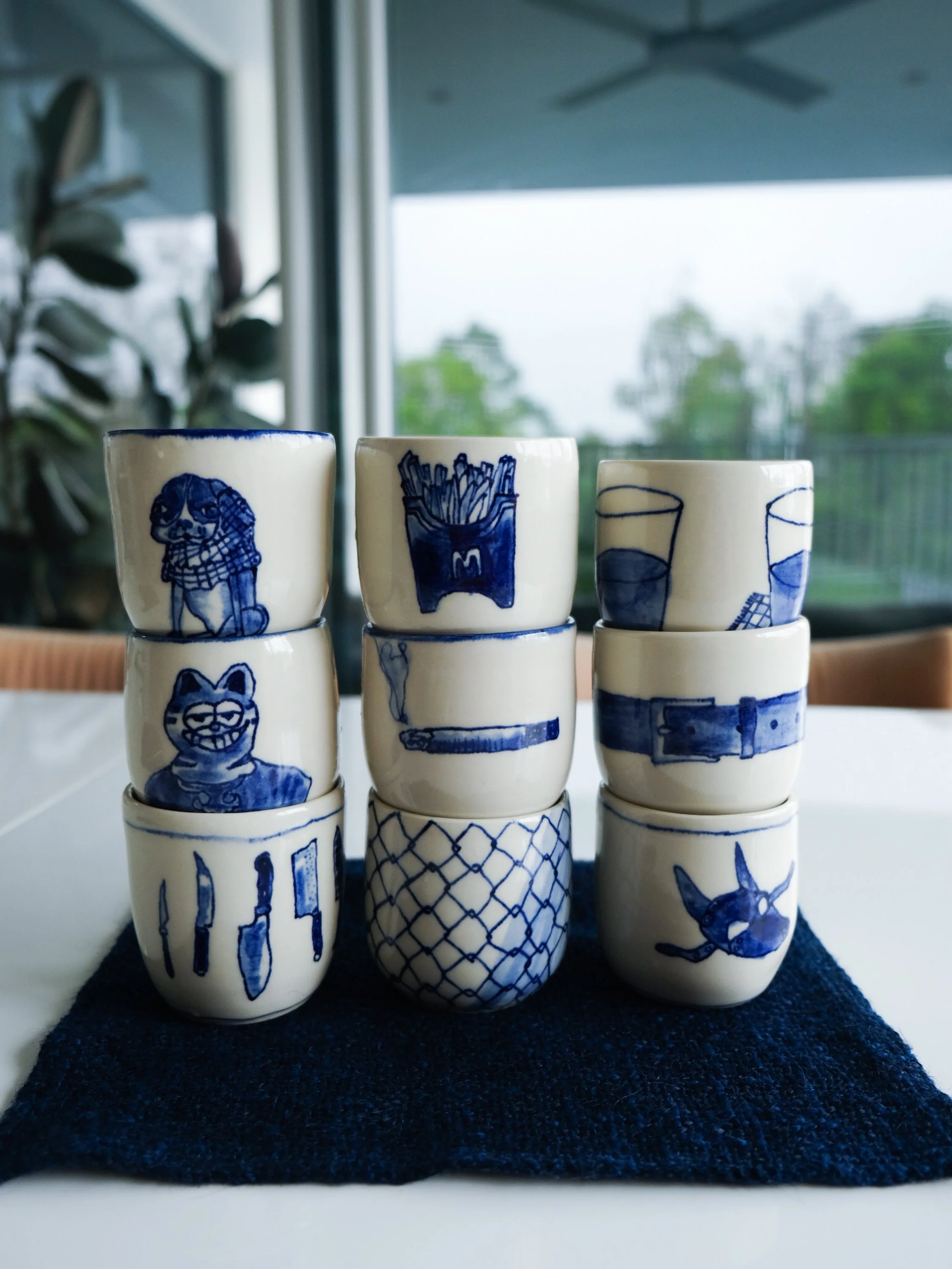 Stacked small white ceramic cups with blue illustrations of animals, objects, and tools, arranged on a dark cloth on a table with a window in the background.