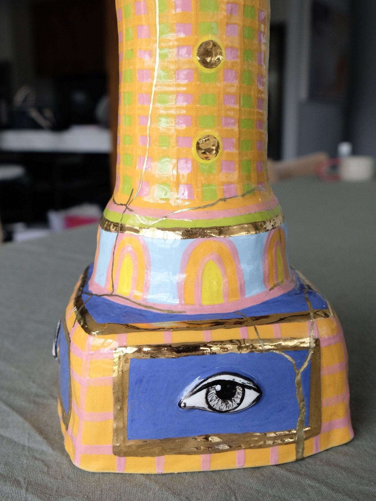 Decorative ceramic sculpture painted with colorful patterns, including an eye on the front, gold accents, and checkered and linear designs.