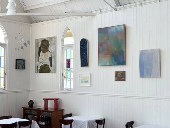Interior of an art gallery with white walls, displaying various colorful paintings, and furniture including tables and chairs.