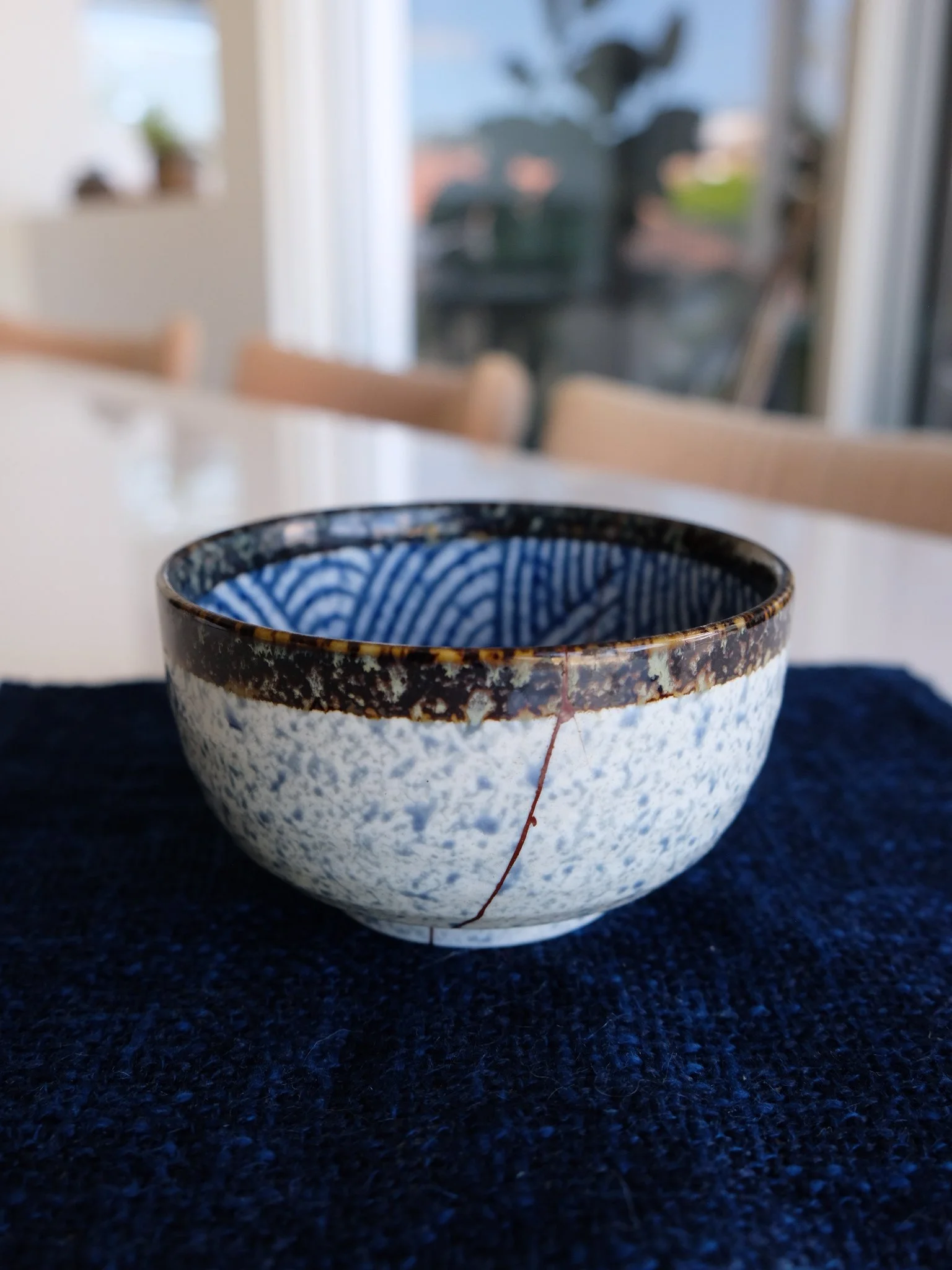 A ceramic bowl with a crack on the side, placed on a dark blue textured cloth, with a blurred background of a room and outdoor view.
