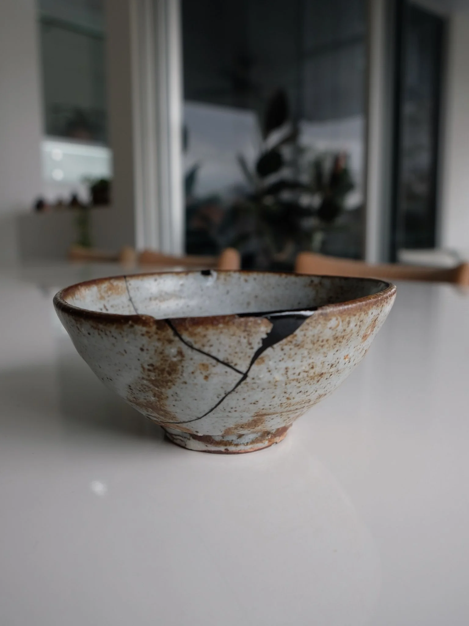 A ceramic bowl with a crack and rust-like speckled glaze on a white surface, indoors with blurred background of furniture and windows. Black urushi kintsugi ceramic repair.