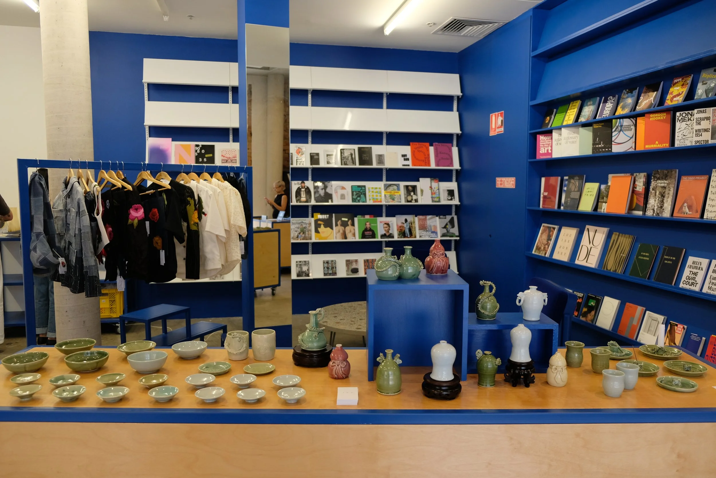 Art and kitchenware display in a bookstore with clothing on a rack and bookshelves in the background.