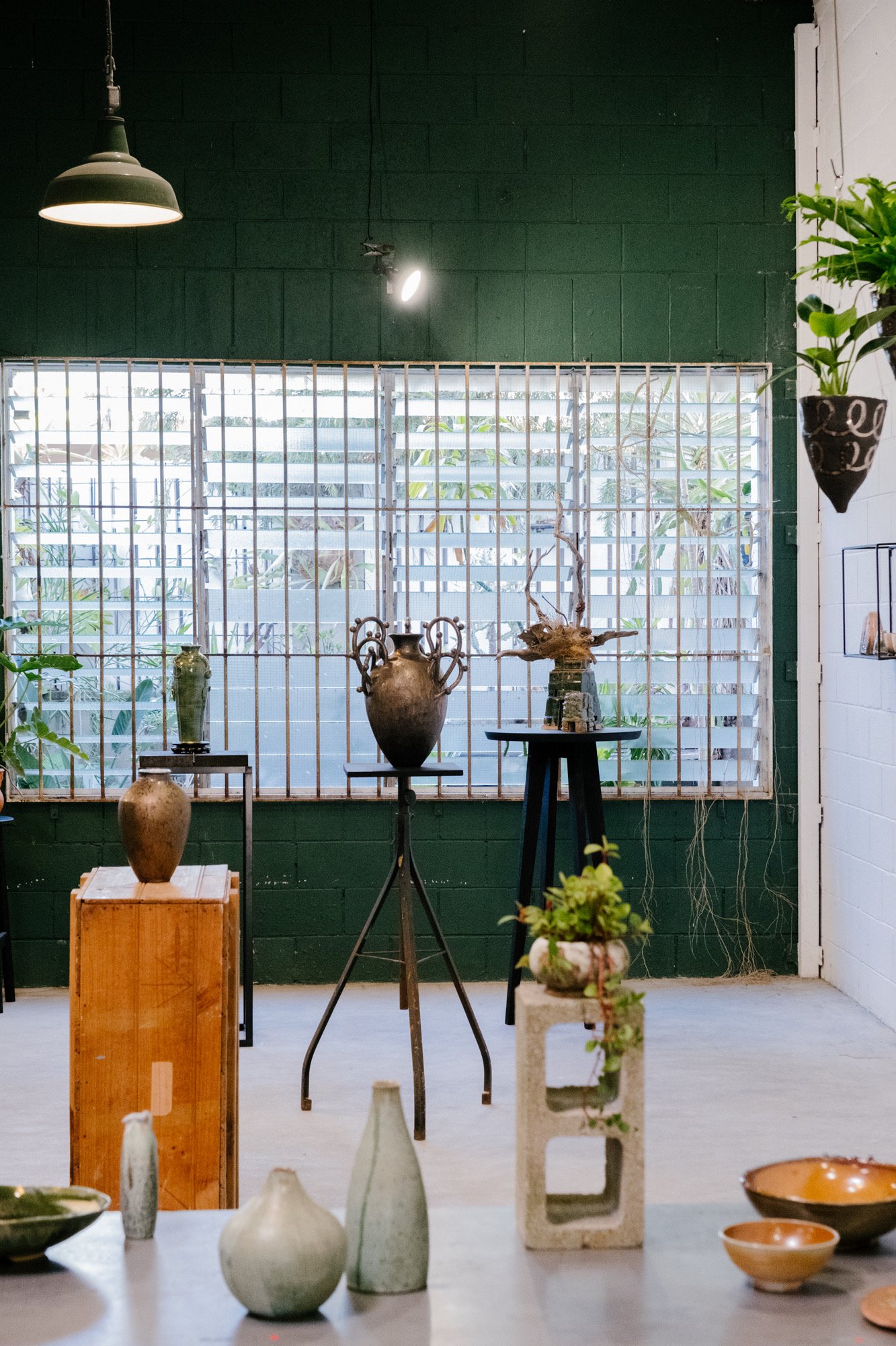 Interior of a ceramics or art studio with ceramic vases and pots displayed on tables and stands, natural light coming through a large window with metal grid, and plants and decorative items visible.