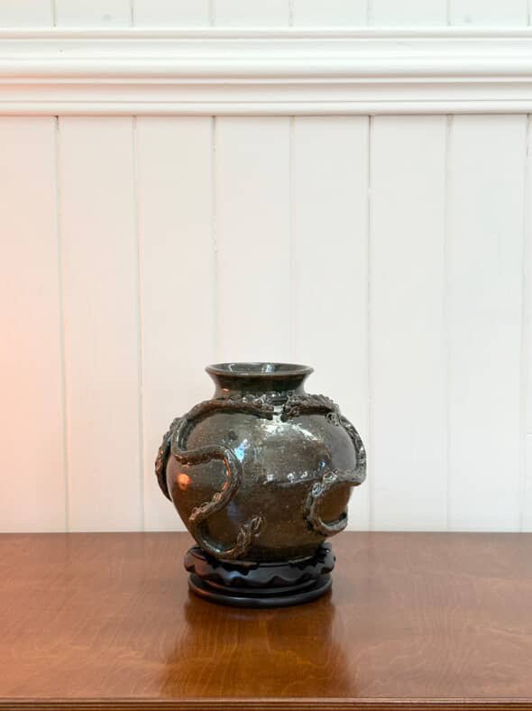 A decorative ceramic vase with ornate handles, placed on a wooden surface against a white paneled wall.